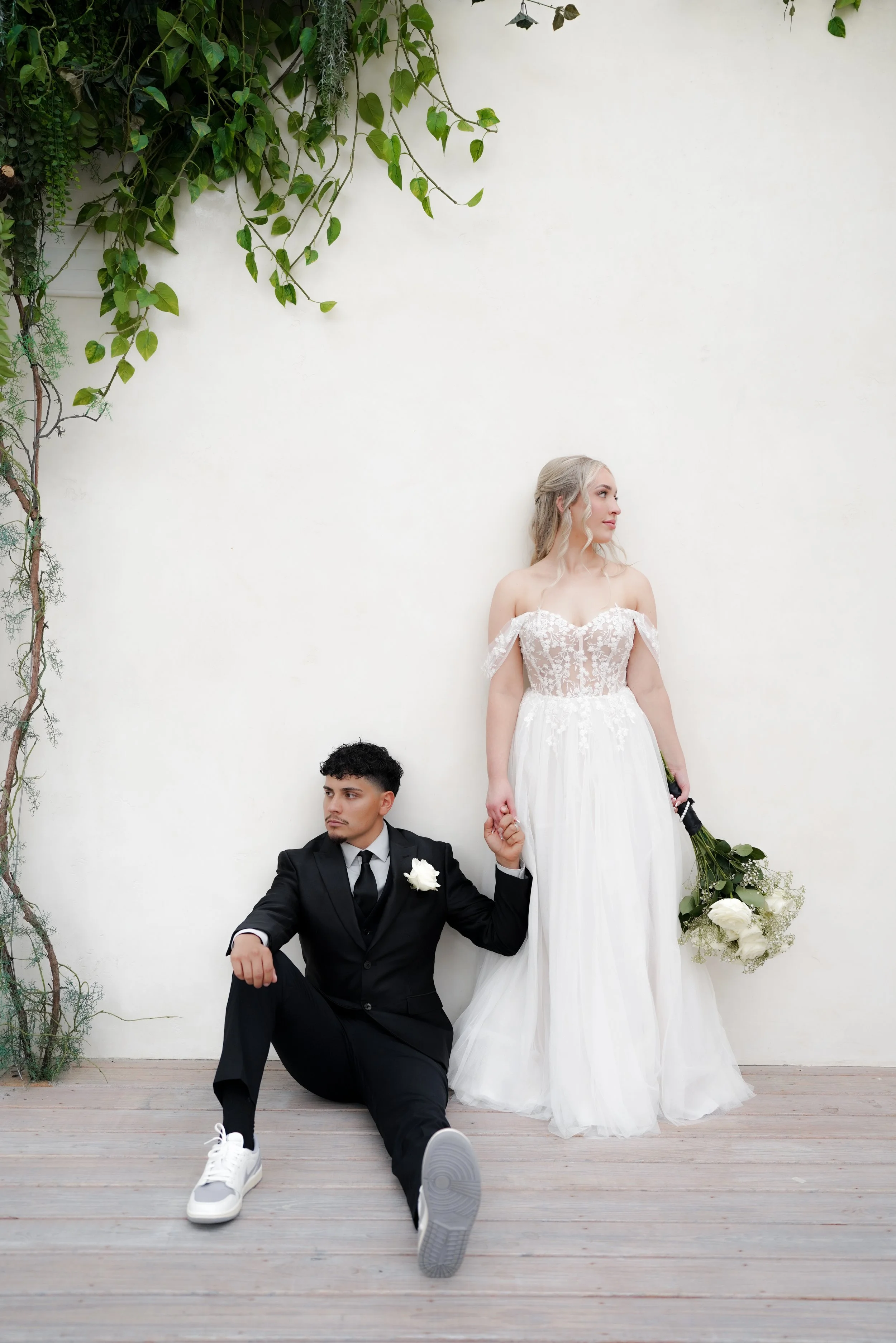 A bride in a white wedding dress stands holding a bouquet of white flowers, and a groom in a black suit sits on the wooden floor, holding her hand, against a plain white wall with green vines hanging from the top left corner.