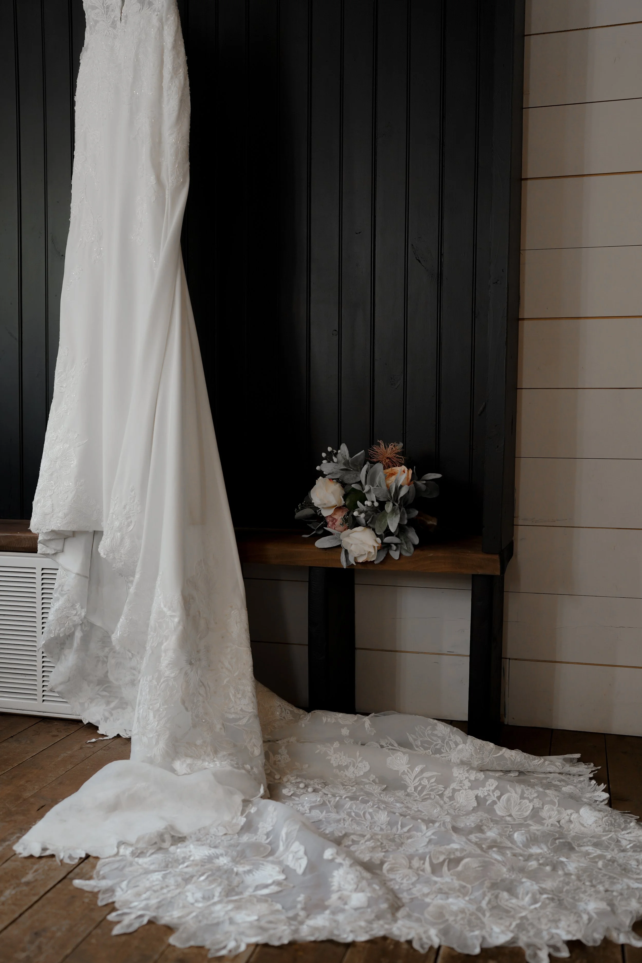 A white wedding dress hanging on a black wooden wall with lace details, beside a bouquet of white and pink flowers resting on a small wooden table.