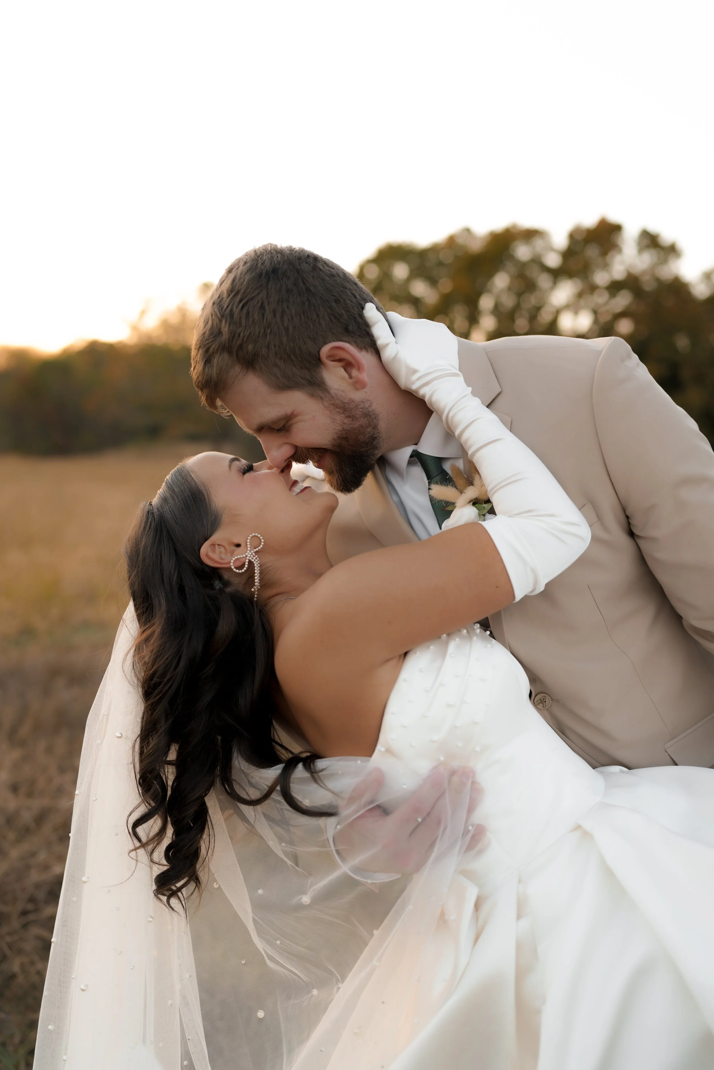 A newlywed couple sharing an intimate moment outdoors at sunset, with the woman in a white wedding gown and veil, and the man in a beige suit, smiling and leaning close.