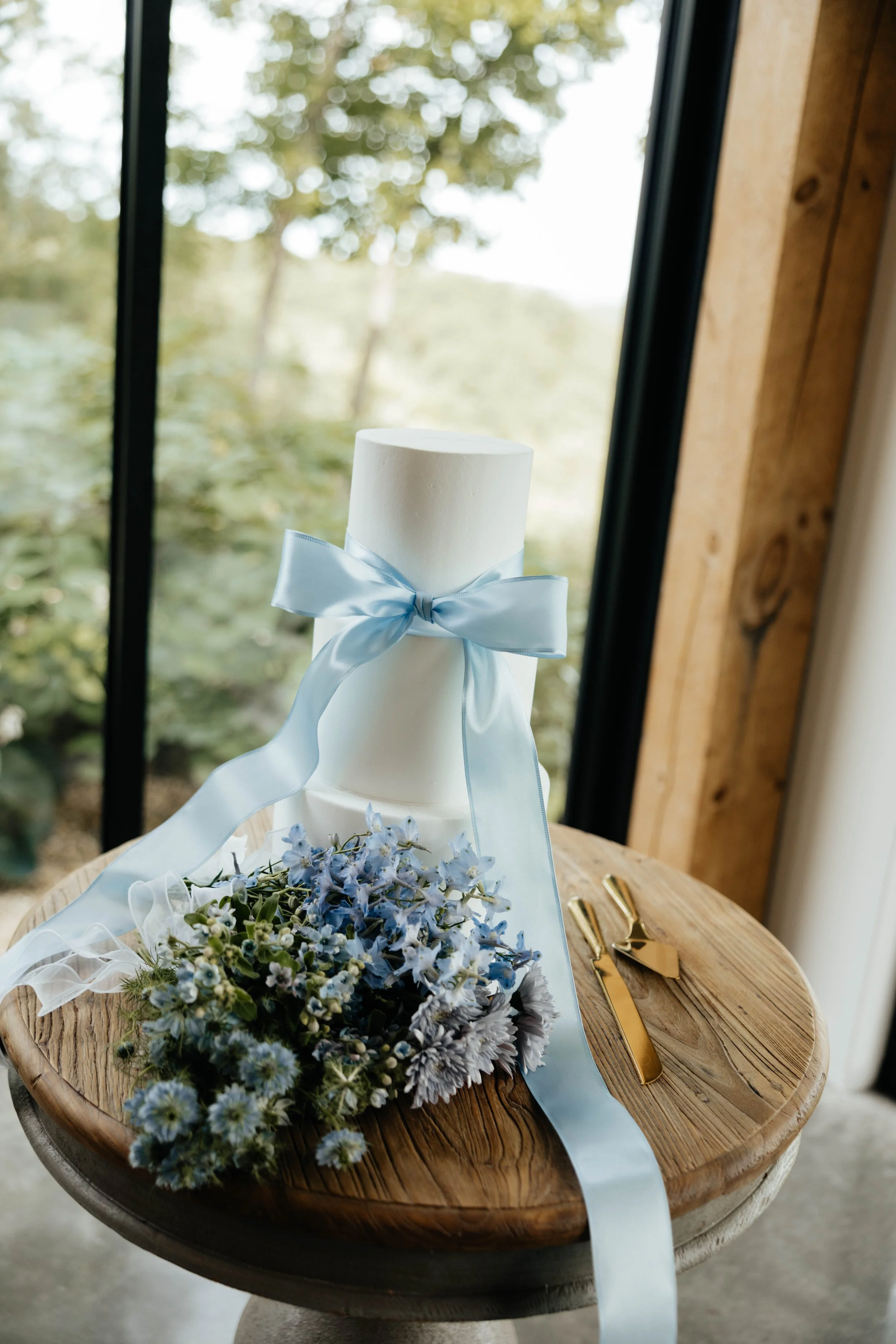 A small round wooden table with a white cylindrical object with a blue ribbon, a bouquet of light blue and white flowers, and gold-colored scissors, set near a window overlooking greenery.