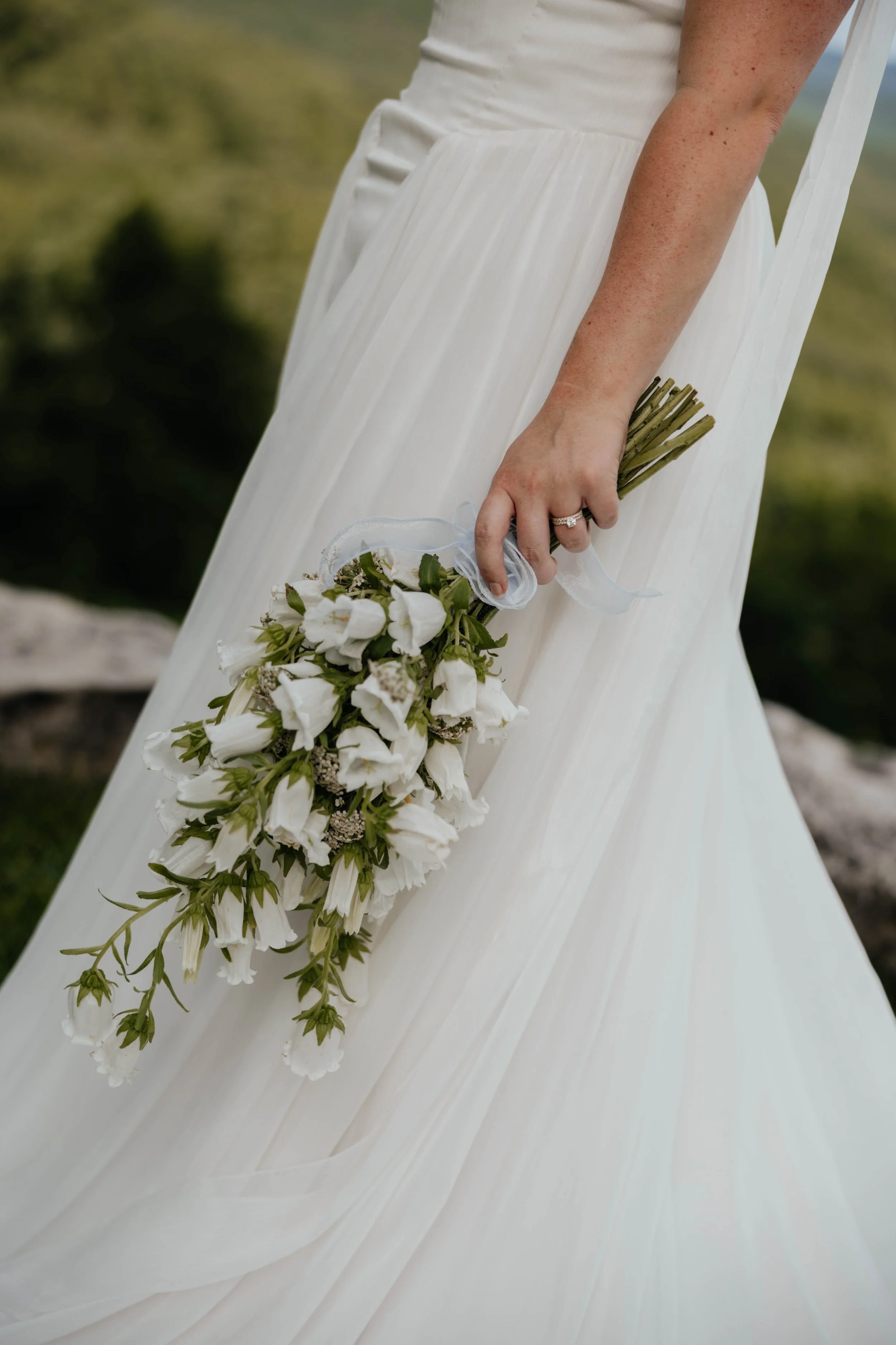 A bride holding a bouquet of white flowers with green stems, wearing a white wedding dress, outdoors with a blurred natural background.