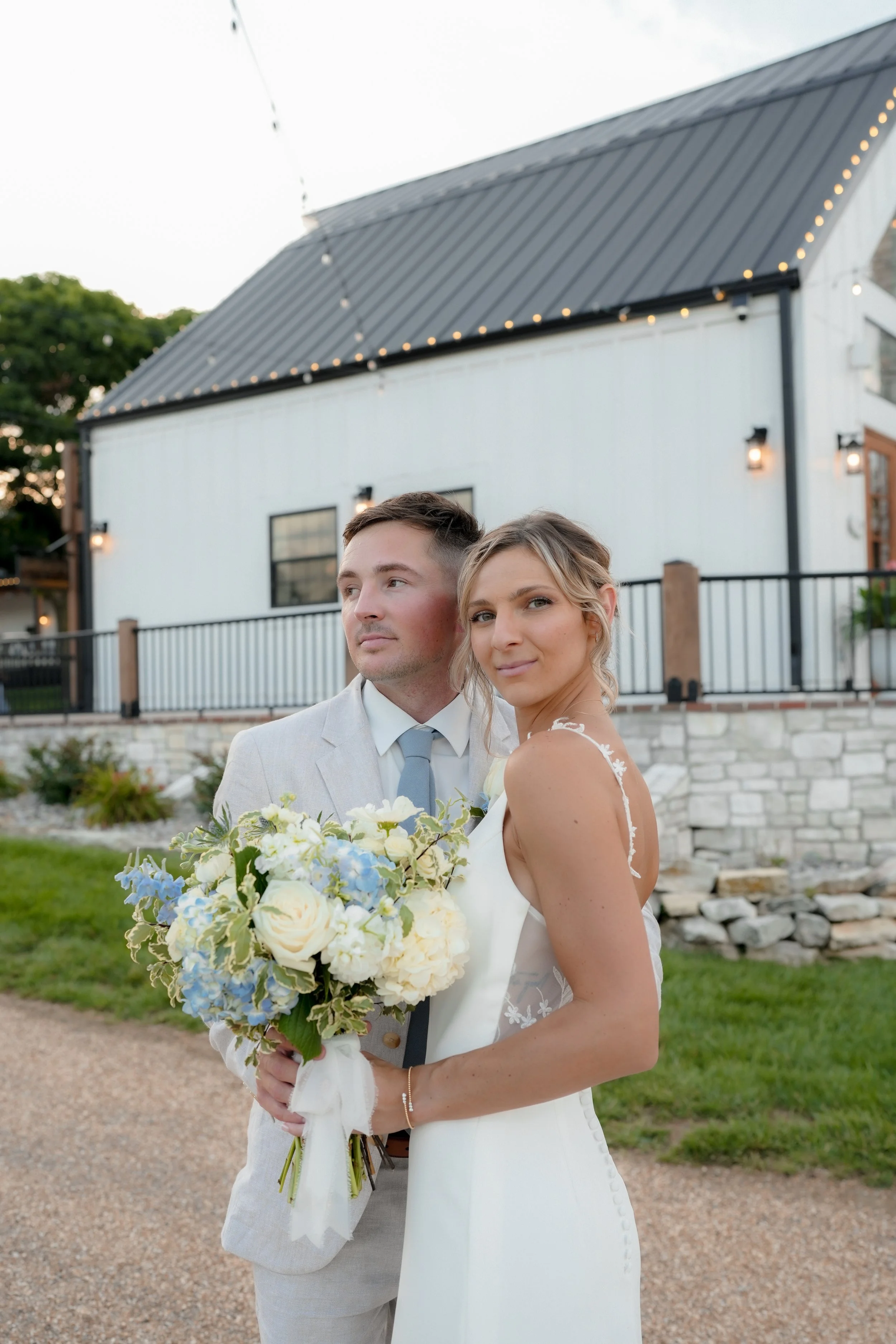 A bride and groom at their wedding outdoors, with the bride holding a bouquet of white and blue flowers, standing in front of a white building with string lights overhead.