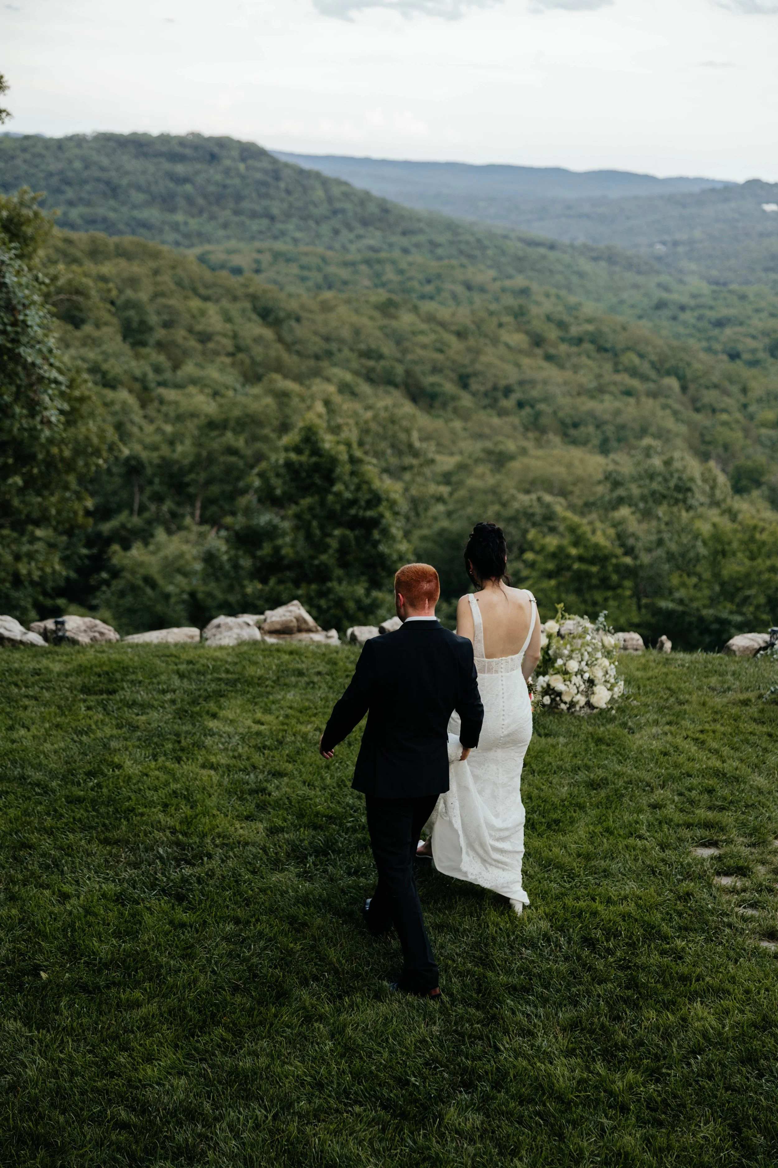 A bride and groom walking away on a grassy hill with a mountain and dense green trees in the background, during a wedding ceremony outdoors.