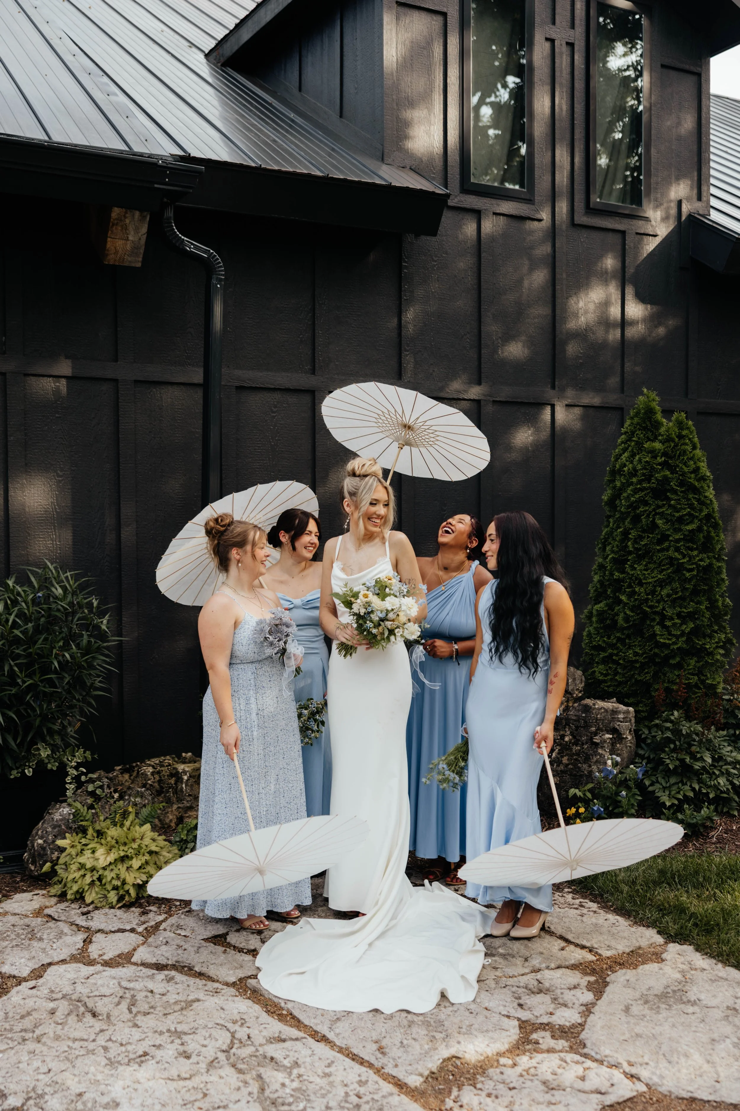 Bride in a white wedding dress surrounded by her bridesmaids in blue dresses, holding white parasols, standing outside in front of a black building with greenery.