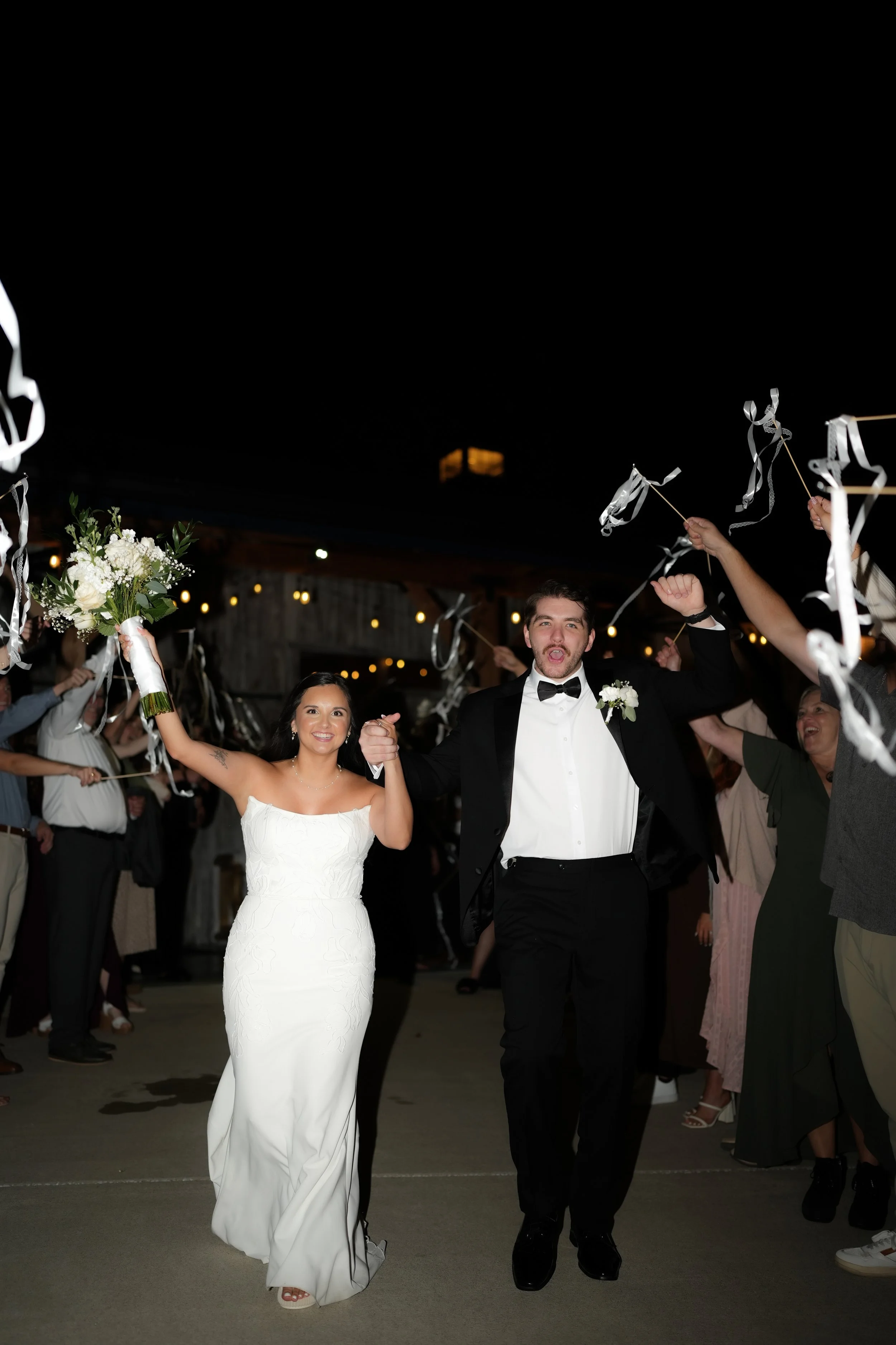 Bride and groom celebrating their wedding, walking hand in hand through a crowd of guests at night, with guests waving ribbons and throwing confetti.