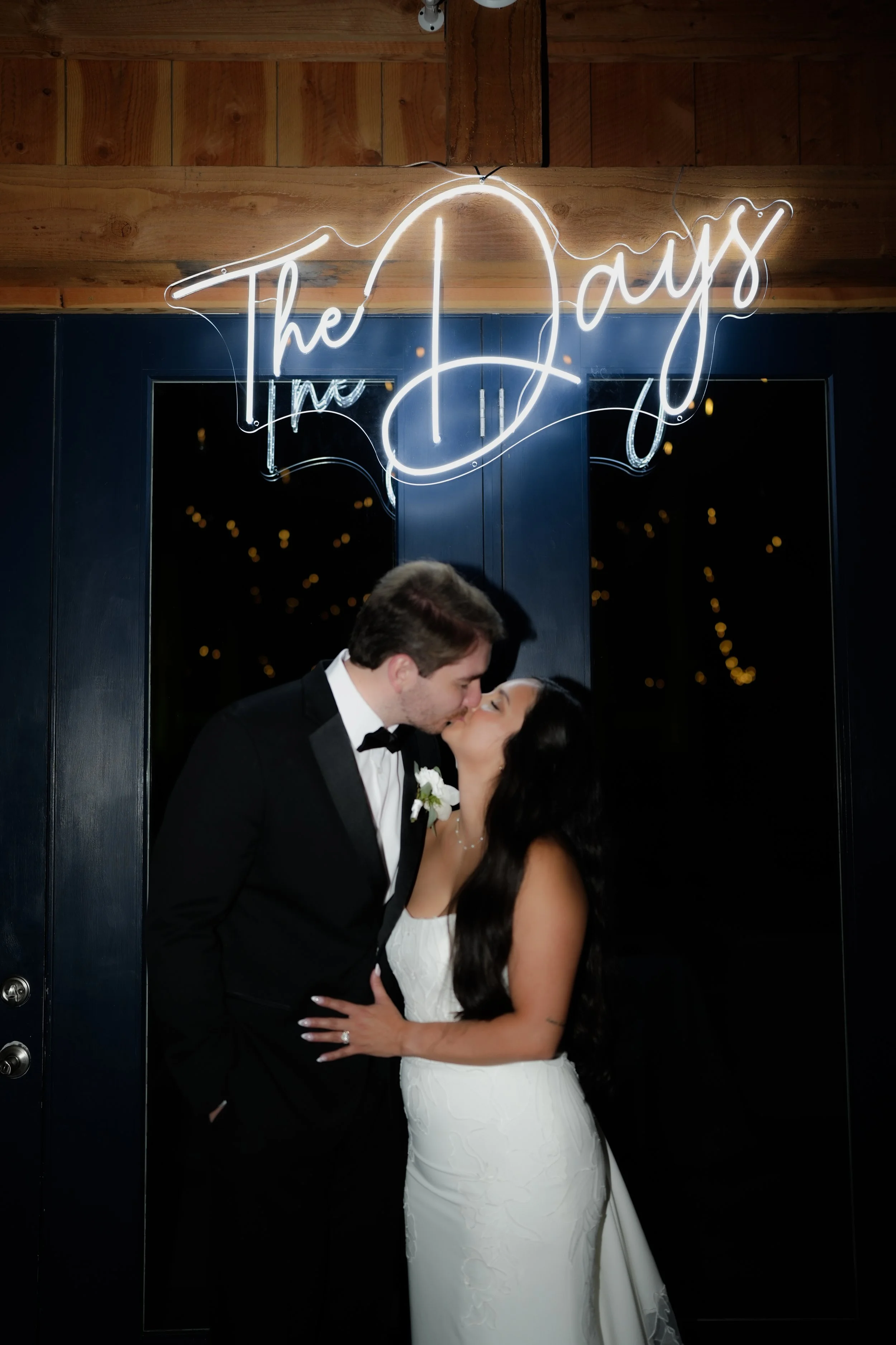A bride and groom sharing a kiss under a neon sign that says 'The Dayz' at their wedding reception.