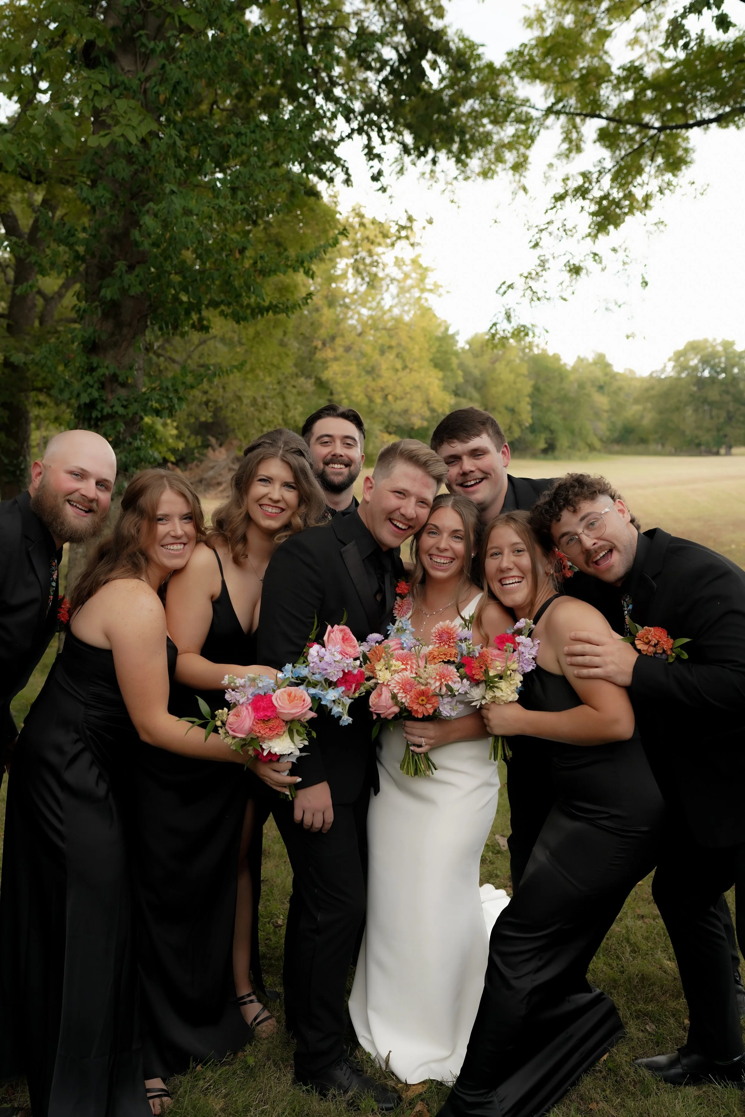 A group of nine people at a wedding outdoors, including a bride, a groom, and seven friends or family members, all smiling and holding colorful bouquets, standing on grass under trees.