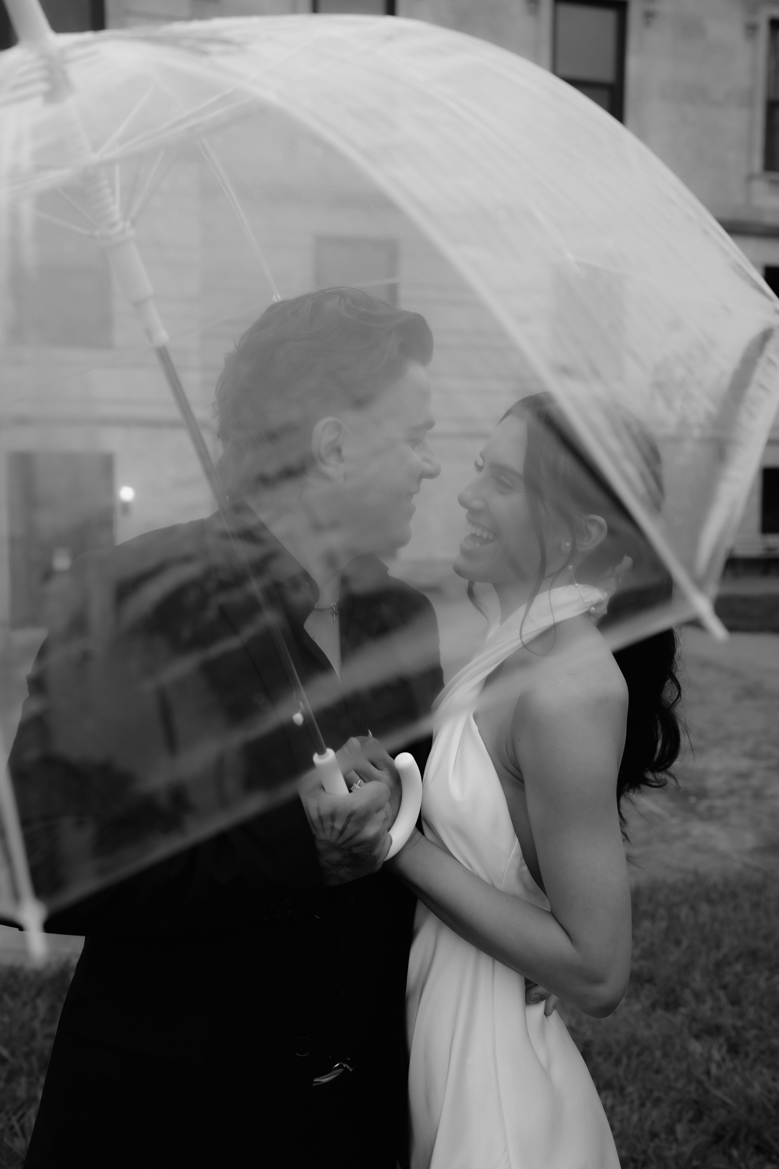 Black and white photo of a man and woman smiling under an umbrella outdoors, with reflections on the umbrella's surface.