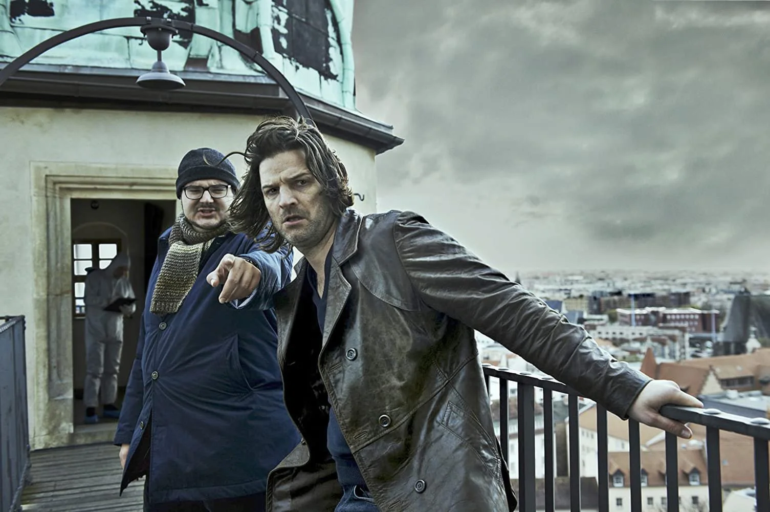 Two men on a rooftop with a cityscape in the background, one pointing at the camera and the other leaning on a railing