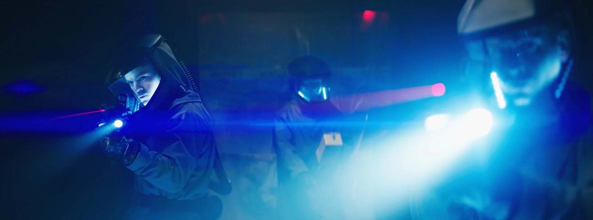 Two individuals wearing helmets and protective gear inside a dark room with bright blue and white lights, possibly exploring or working in a high-tech environment.