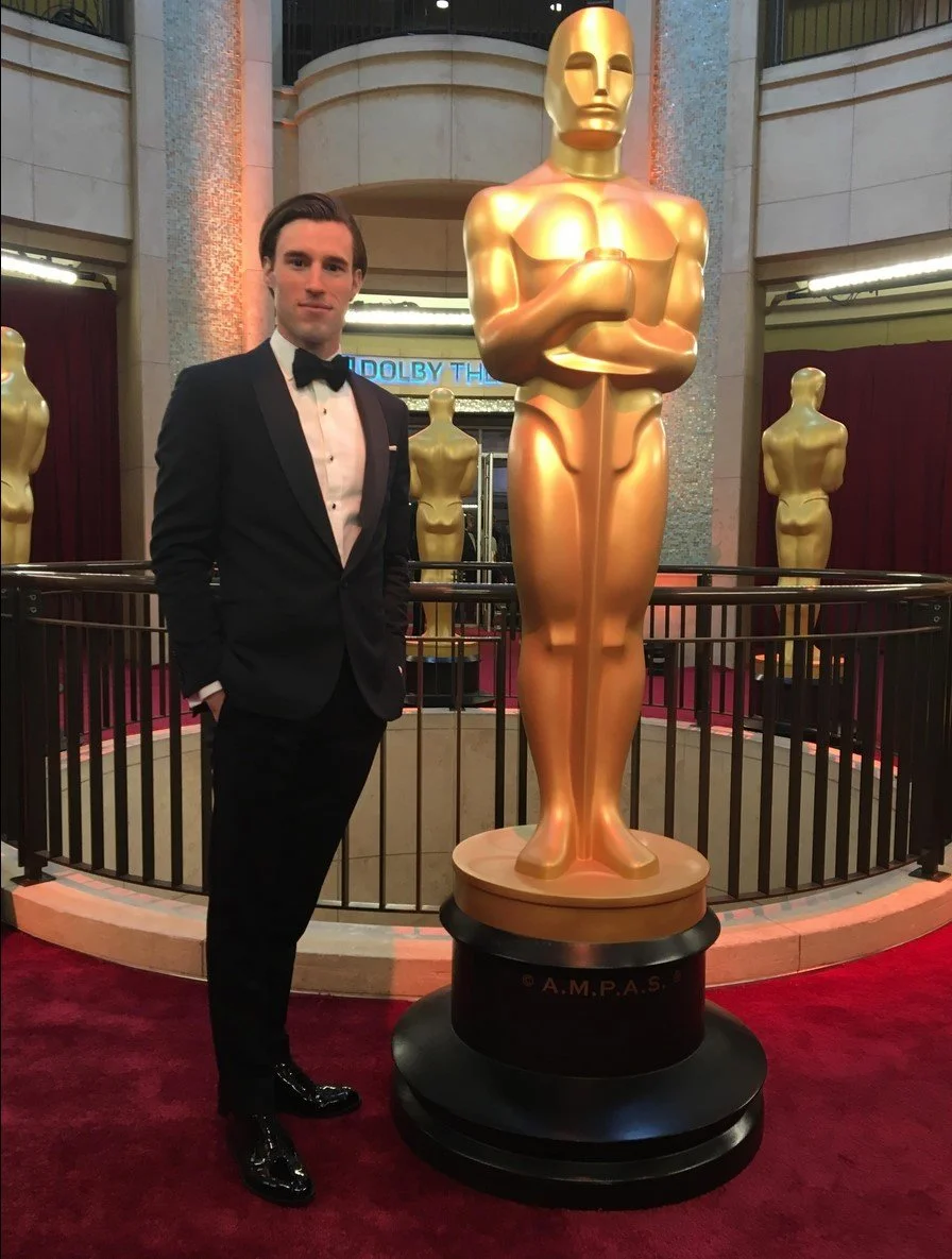Man in tuxedo standing next to a large golden Oscar statue.