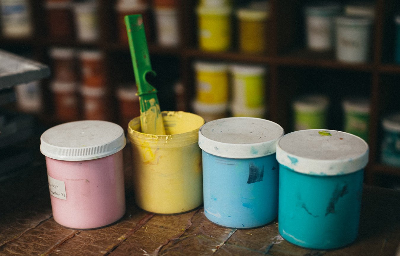 Four small containers of paint in pink, yellow, and two shades of blue with a paintbrush inside the yellow container on a wooden surface, with shelves of more paint containers in the background.