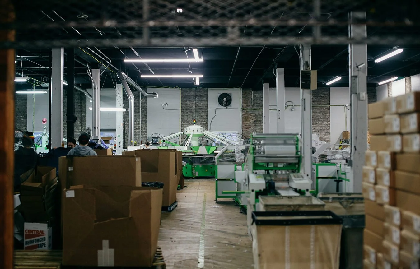 Inside a factory or warehouse with workers and machinery, including packing stations and stacks of boxes.