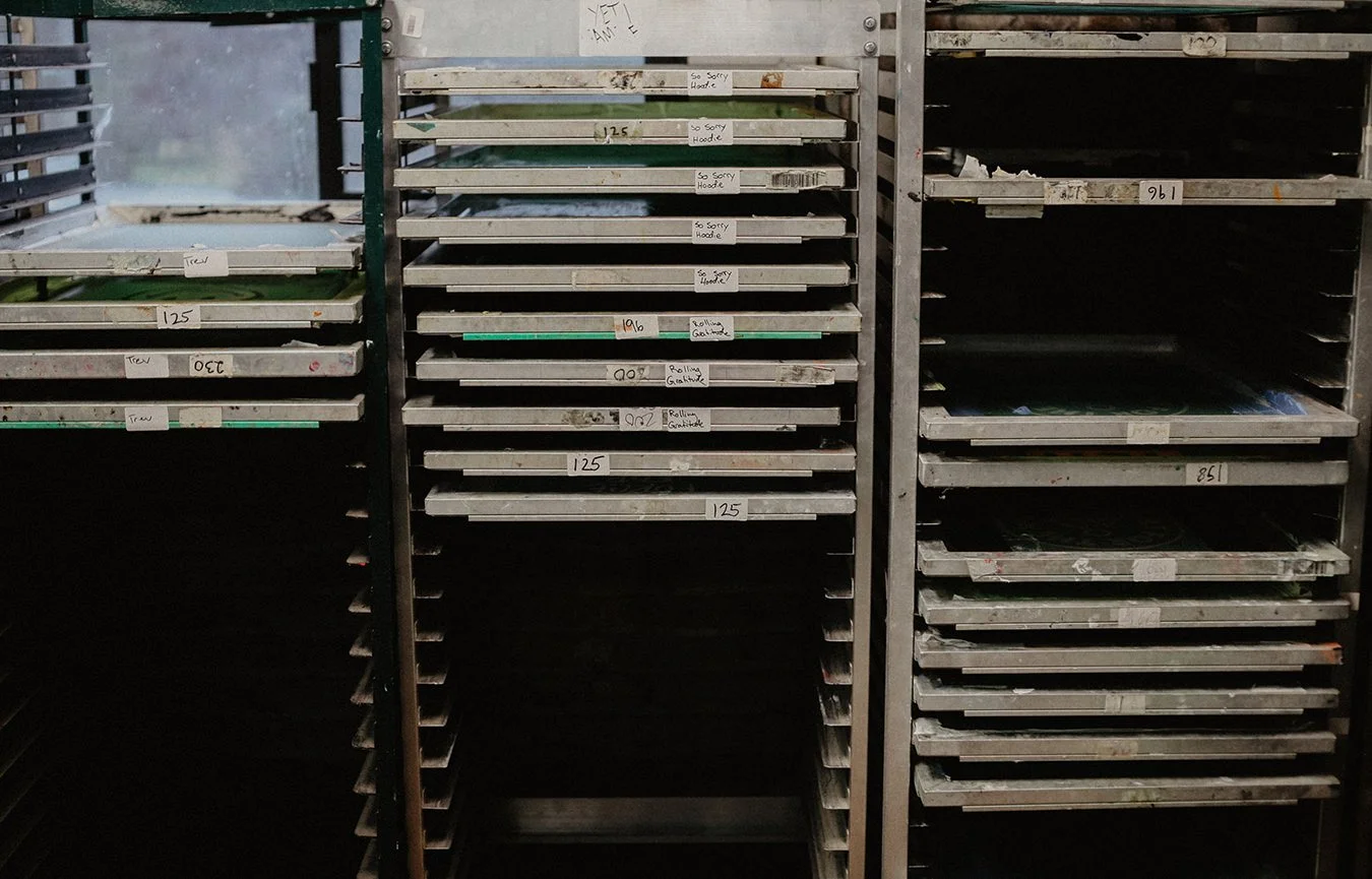 Metal shelving units with empty and partially filled trays labeled with handwritten tags and numbers in a storage or industrial environment.