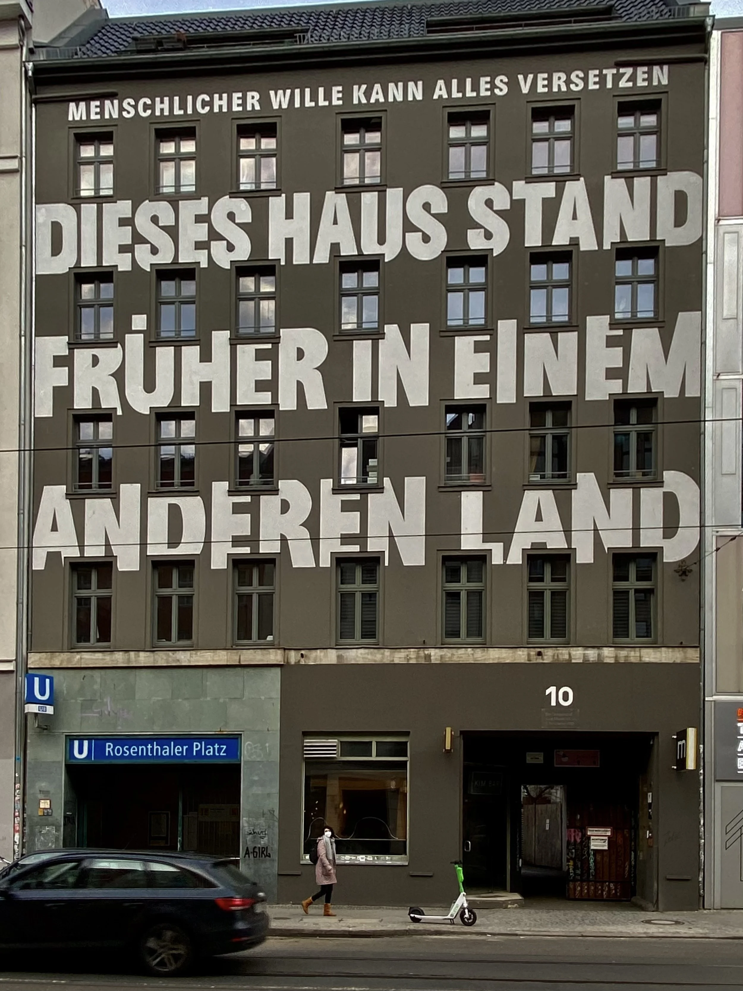 Ein mehrstöckiges Gebäude mit einer großen Wandwerbung, die einen philosophischen Gedanken in deutscher Sprache zeigt. Vor dem Gebäude fahren Autos, und es ist eine Person mit einem Rucksack und einer Maske zu sehen.