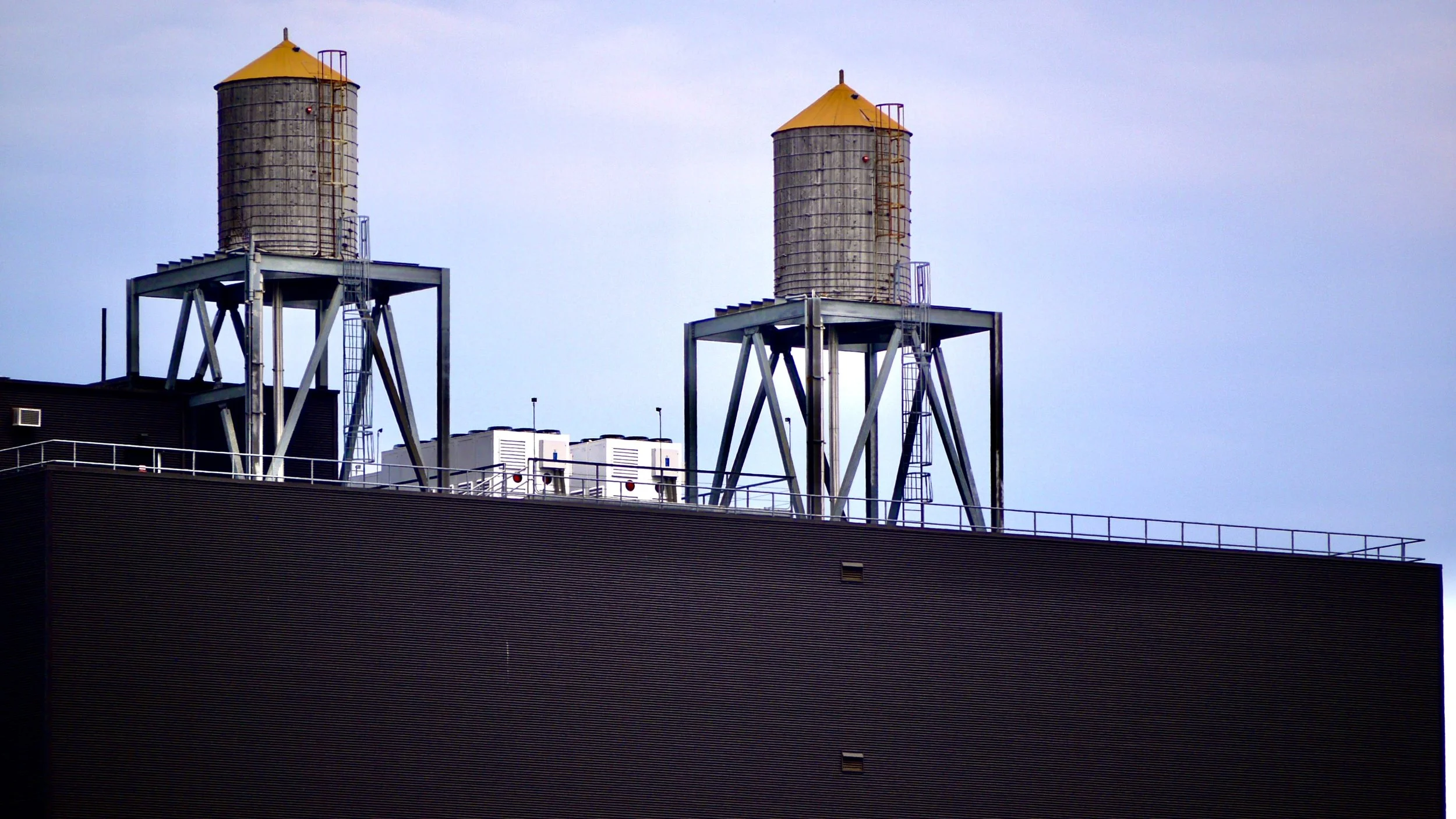 Zwei Wasserreservoirs auf dem Dach eines Gebäudes, umgeben von einer schwarzen Fassade und einem separates Geländer, mit einem klaren blauen Himmel im Hintergrund.