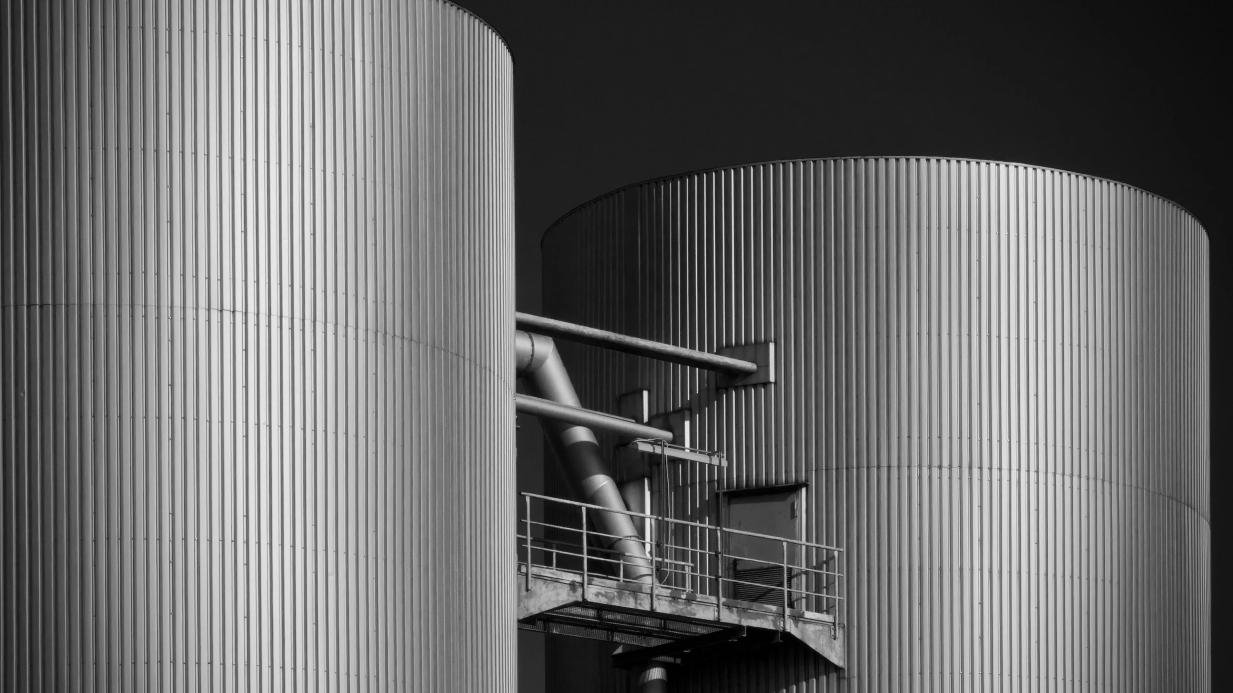 Schwarz-Weiß-Foto von zwei runden industriellen Lagertanks mit vertikaler Riffelung, verbunden durch Rohre und Geländer, unter dunklem Himmel.