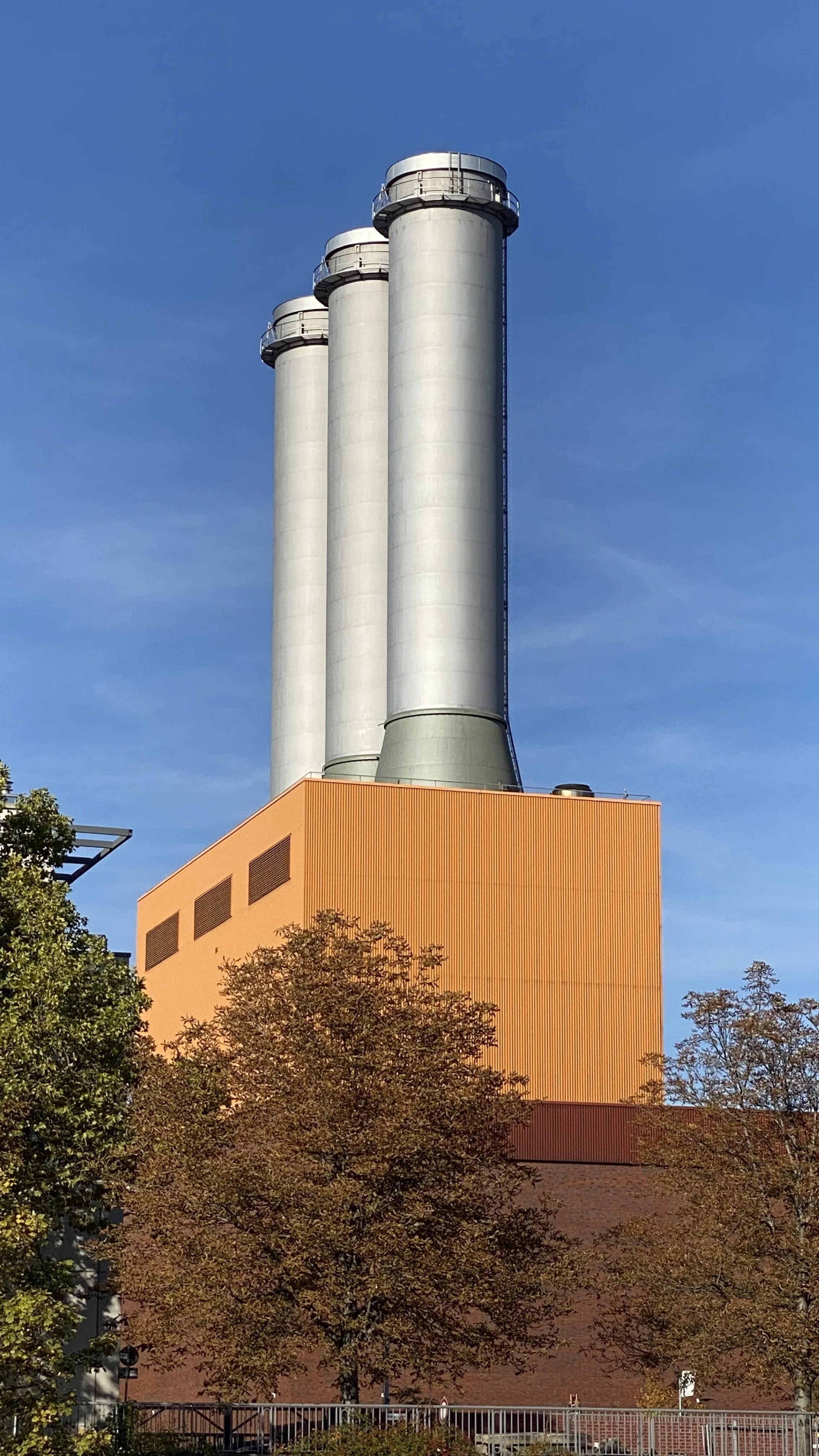 Industrialanlage mit drei hohen silbernen Schornsteinen vor einem blauen Himmel, umgeben von Bäumen mit herbstlichen Blättern.