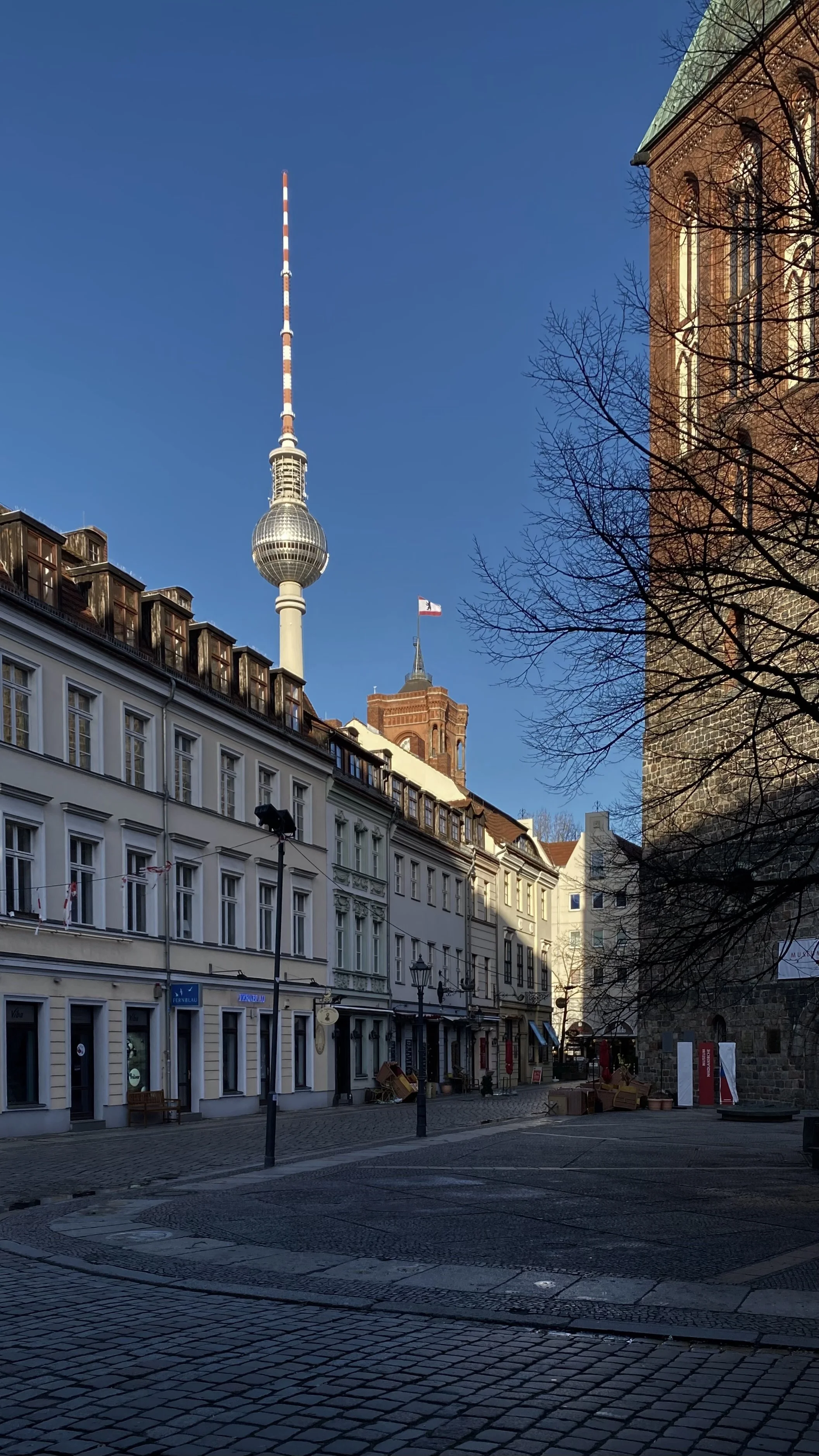 Blick auf eine Stadtstraße mit historischen Gebäuden, dem Fernsehturm und der Universität Berlin im Hintergrund bei Sonnenschein.