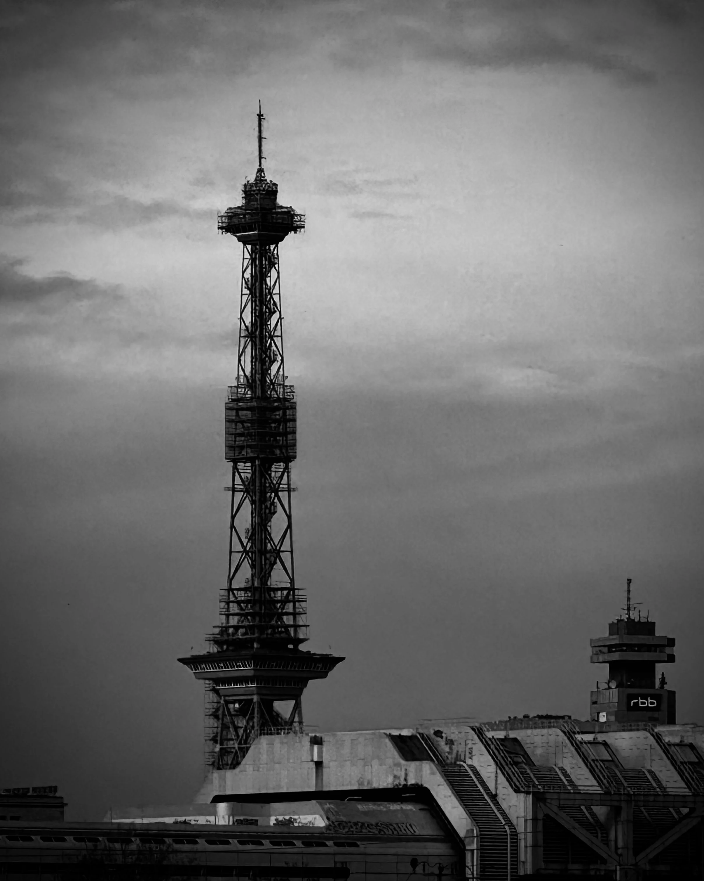 Schwarz-weiß-Bild mit einem hohen Fernsehturm und einem Gebäudeteil im Vordergrund, Himmel im Hintergrund.