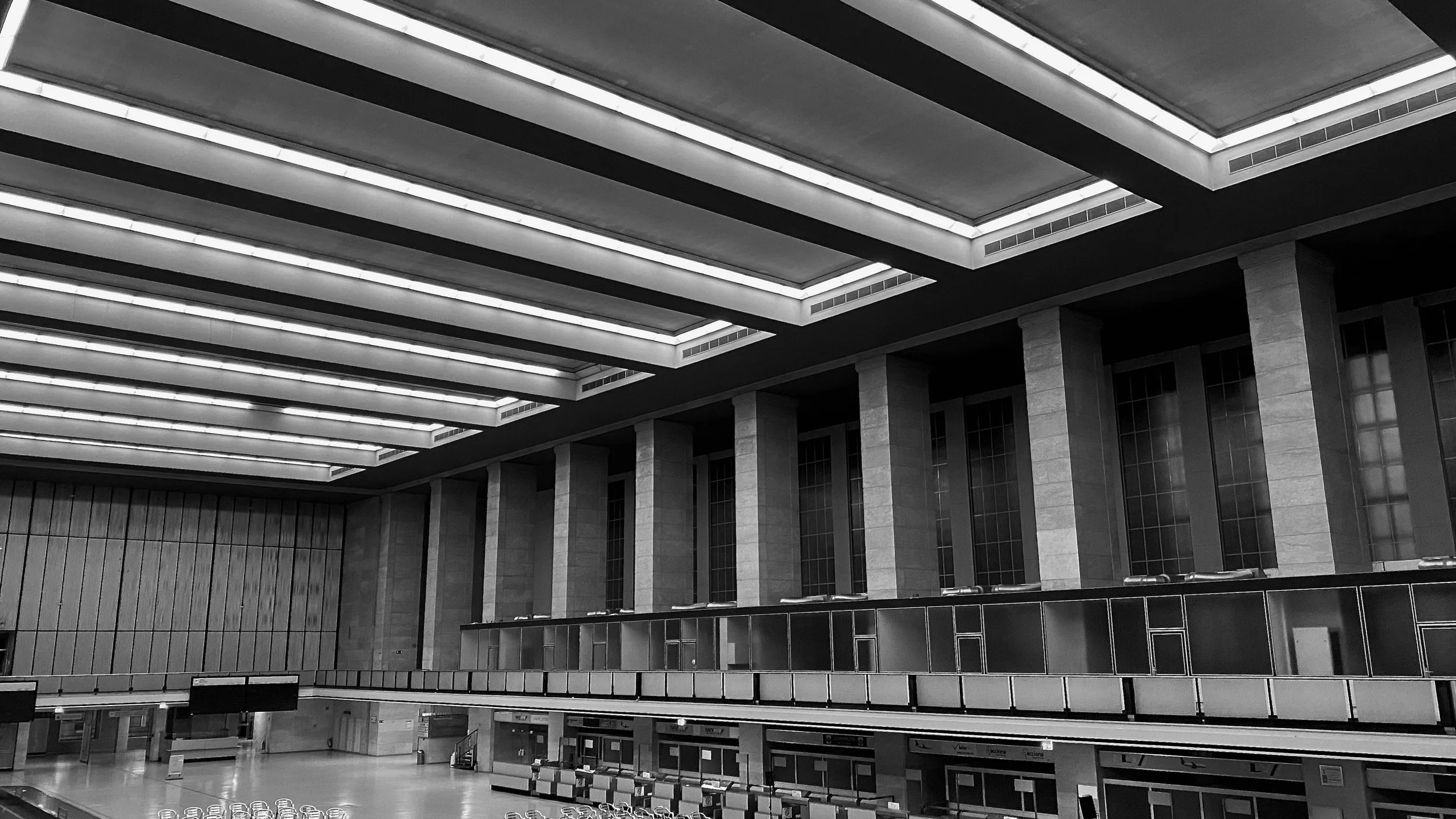 Schwarz-weiß Foto eines modernen, leeren Flughafenterminals mit hohen Decken, großen Fenstern und Sitzbereichen.