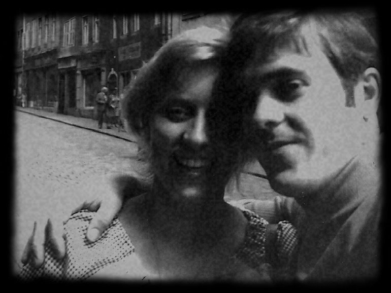 Ein Schwarz-Weiß-Foto von zwei Personen, die eng beieinander stehen und sich umarmen, auf einer Stadtstraße mit Gebäuden im Hintergrund.