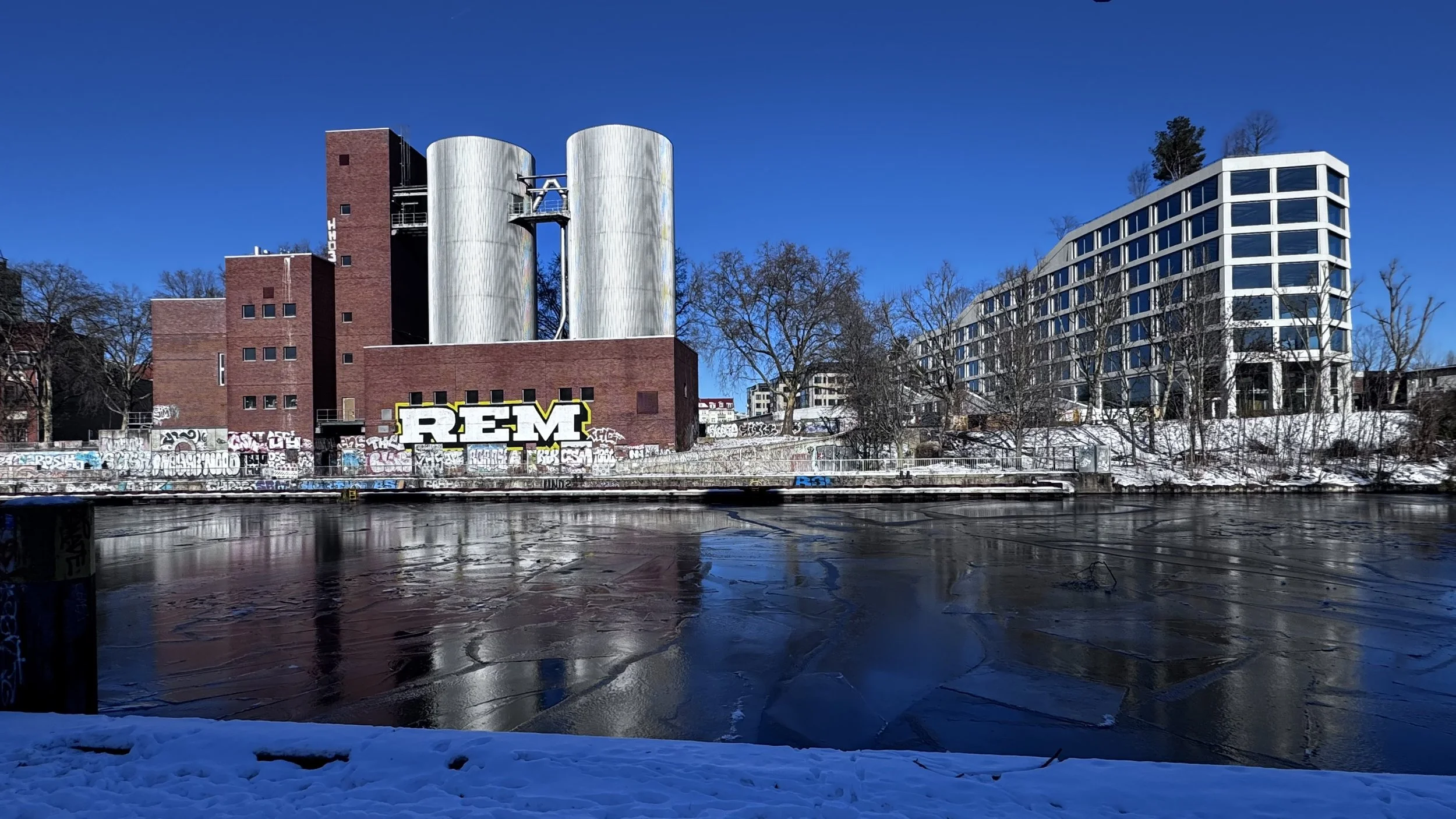 Städtisches Gebiet im Winter mit gefrorenem Fluss, Graffiti an Wänden, moderne weiße Gebäude und ein Industriebau mit Metalltanks unter klarem blauen Himmel.