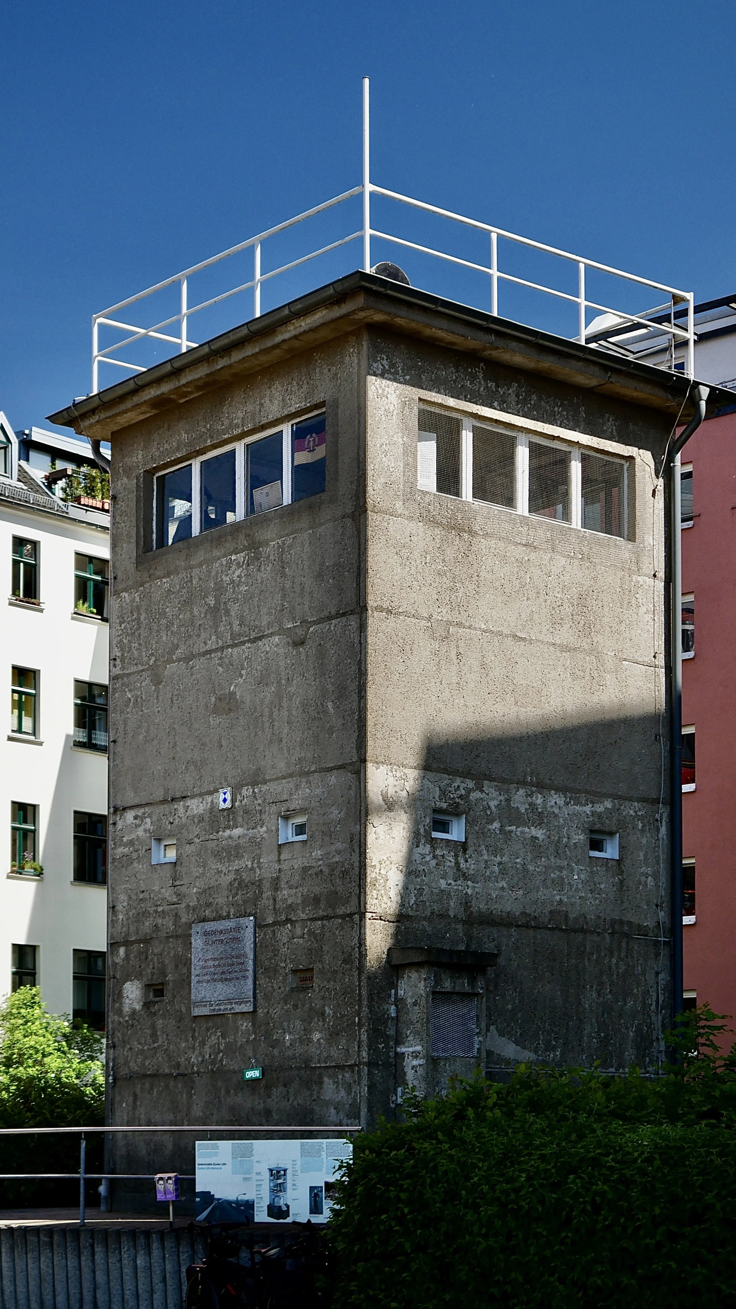 Ein alter, grauer Betonturm mit kleinen Fenstern und einem Geländer auf dem Dach, umgeben von modernen Gebäuden.