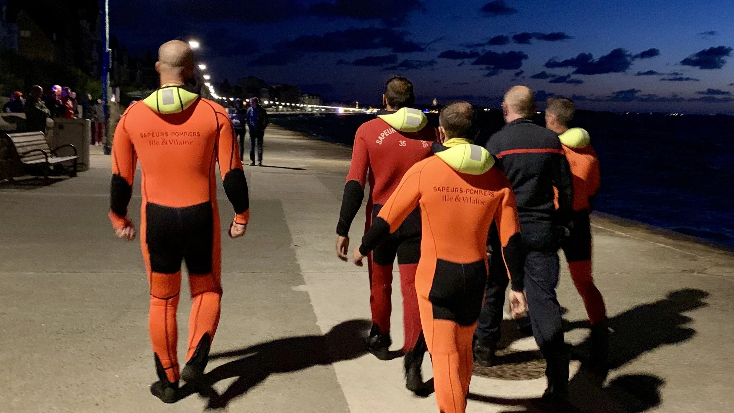 Rettungssanitäter der Feuerwehr Ile et Vilaine im orange-schwarzen Einsatzanzug am Strand bei Nacht, der Blick ist nach vorne gerichtet.
