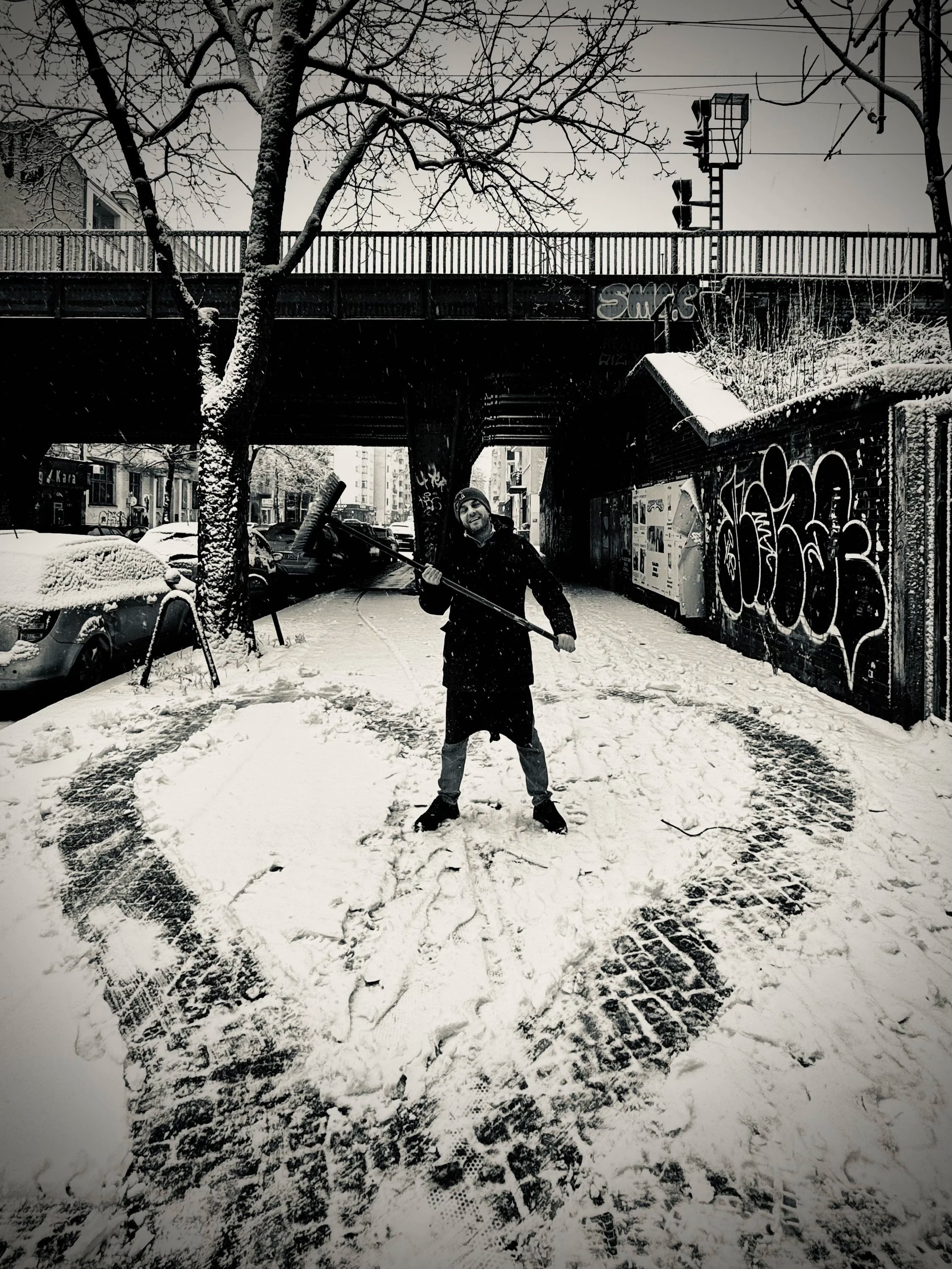 Ein Mann steht im Schnee unter einer Brücke, hält eine Schneeschaufel und lächelt. Der Boden ist mit Fußgängerspurten bedeckt, umgeben von grauen, mit Graffiti besprühten Wänden, parkenden Autos und einer Schneelandschaft.