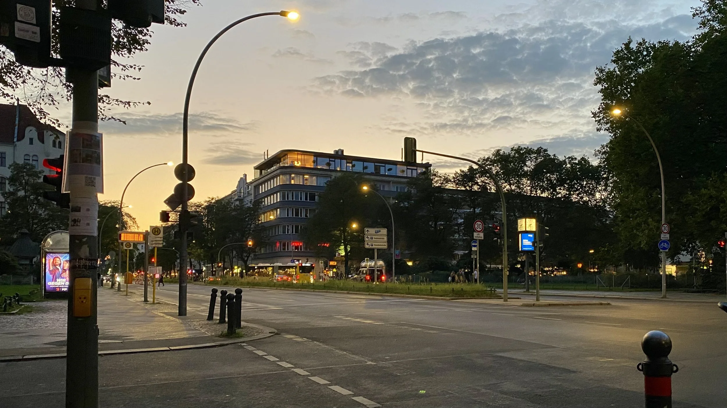 Stadtszene bei Sonnenuntergang mit Straßenlaternen, Verkehrsampeln, Gebäuden und Bäumen.