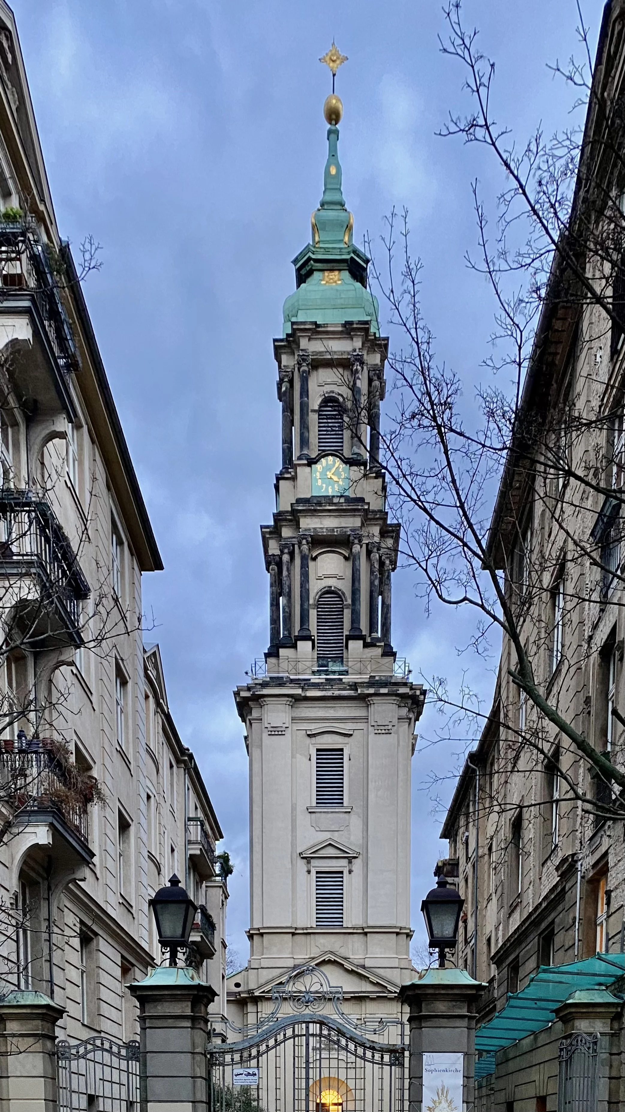 Ansicht eines Barockhauptturms einer Kirche mit Uhr und goldener Spitze, umgeben von alten Gebäuden und Bäumen.