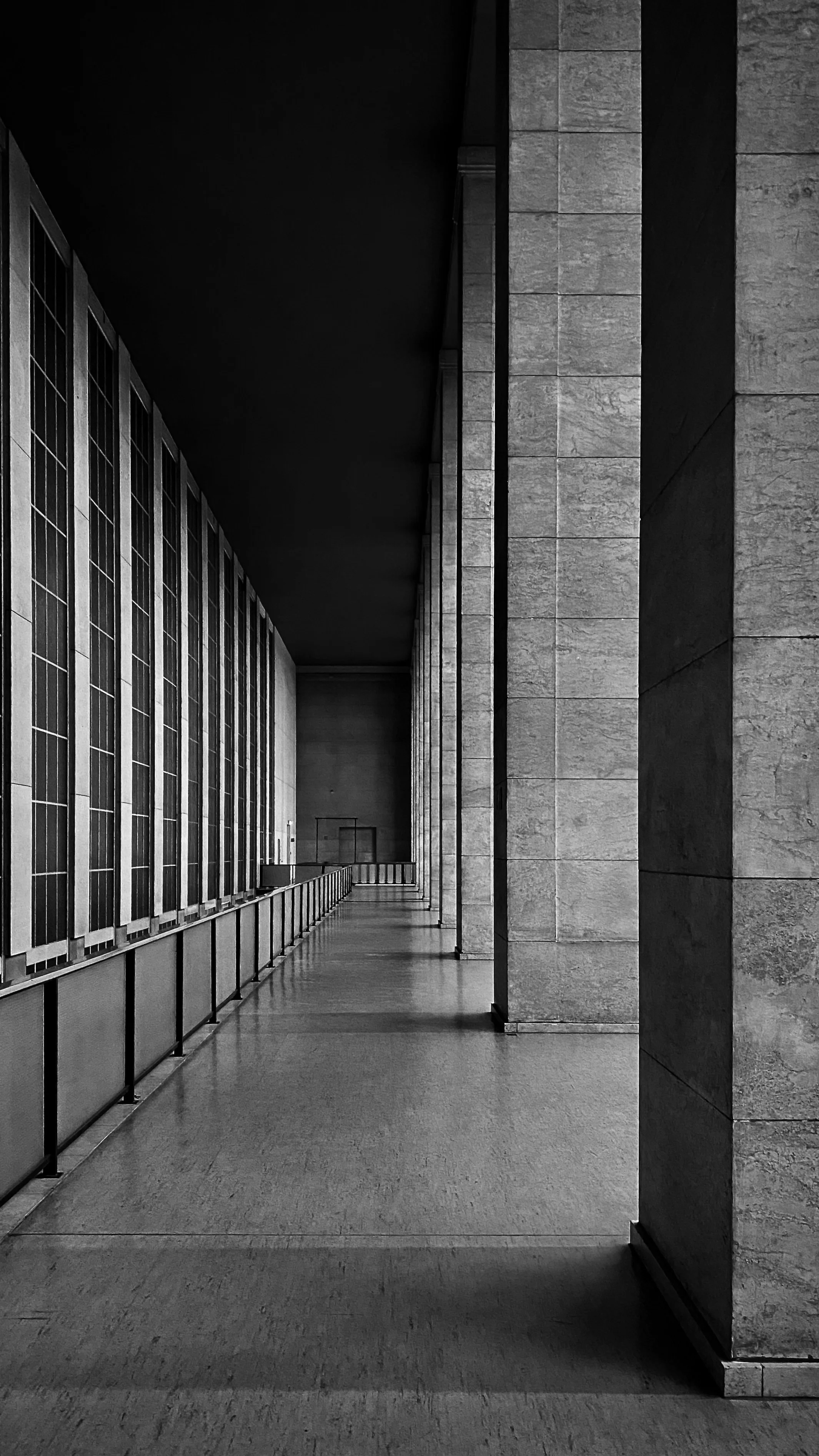 Schwarz-weiß-Foto eines langen Flurs mit großen Fenstern auf der linken Seite und mehreren großen Steinsäulen auf der rechten Seite.