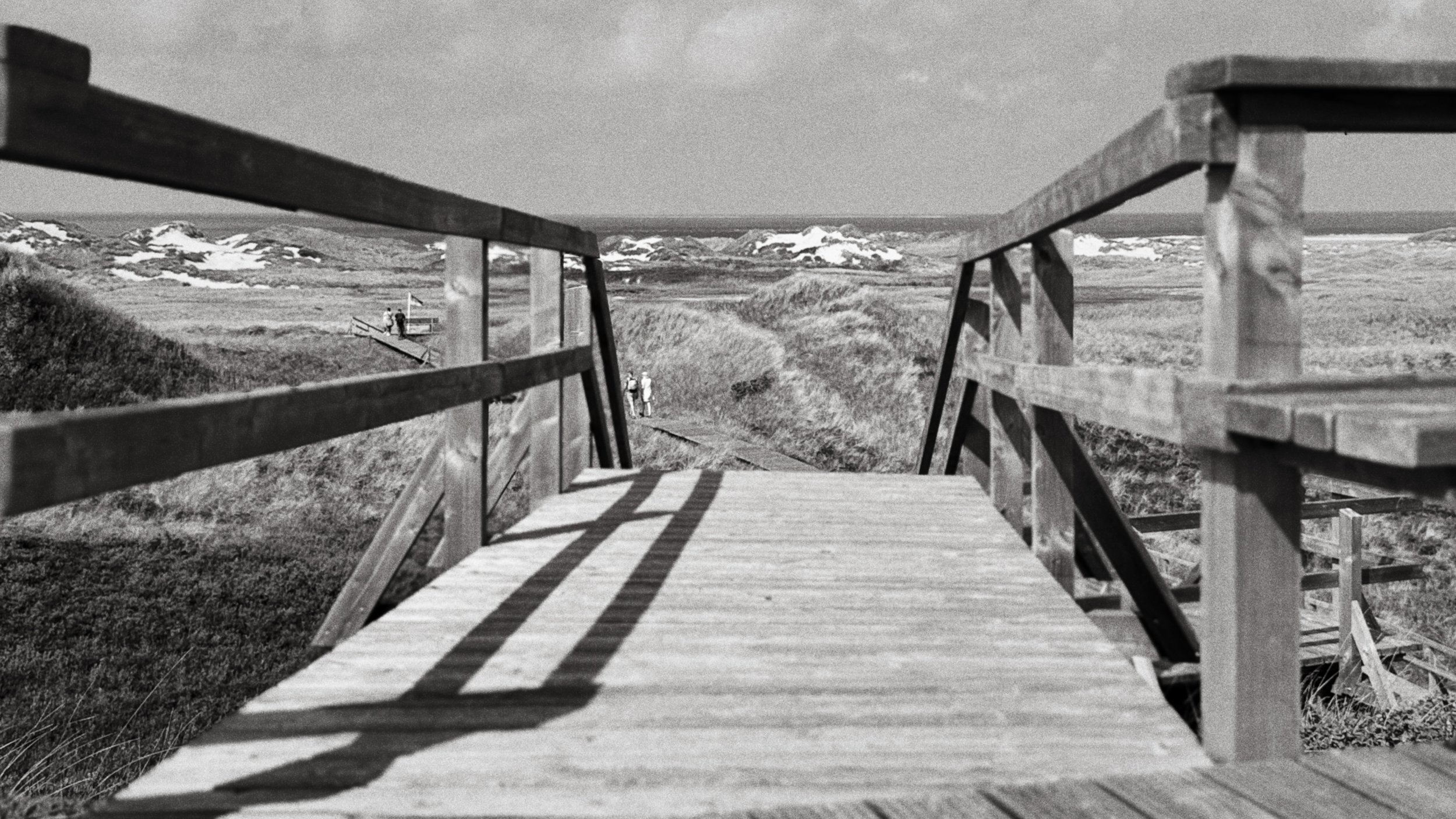 Blick von einer Holzbrücke auf eine ländliche Landschaft mit Wiesen und Schneefeldern im Hintergrund, einige Menschen gehen auf dem Weg.