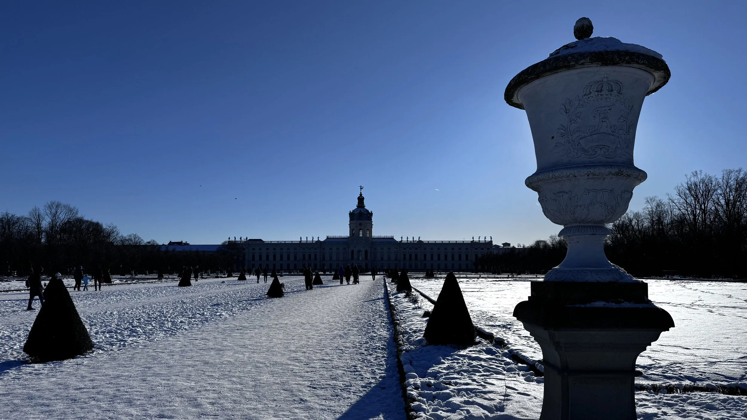 Ein Winterbild mit Schnee bedeckter zeitloser Garten, im Hintergrund ein barockes Gebäude mit einer Uhrturm. Im Vordergrund eine große dekorative Steingutschale und kleine konische Bäume. Klarer blauer Himmel, viele Menschen im Garten.