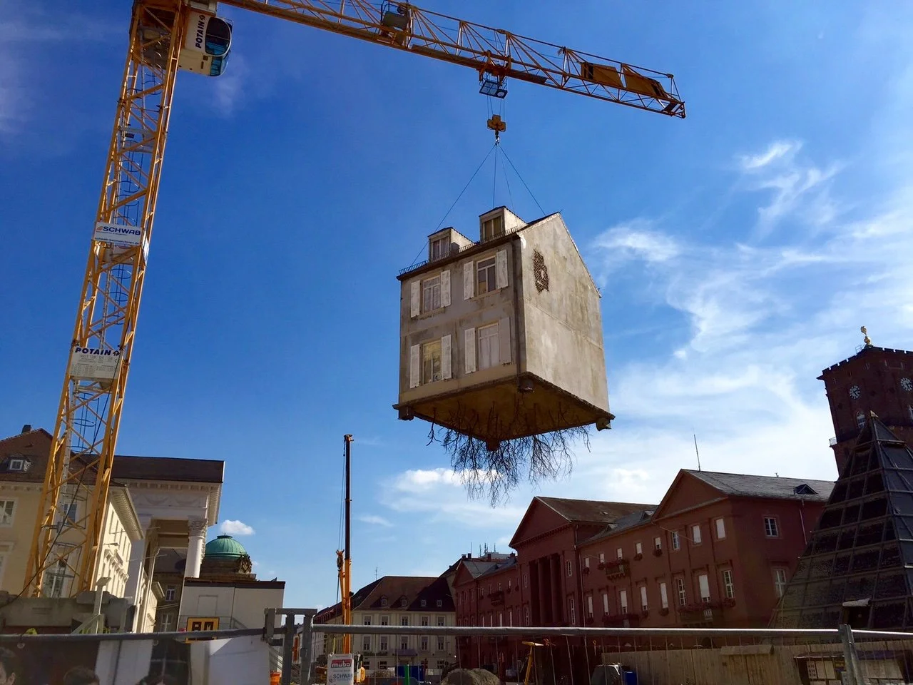 Ein Kran hebt ein altes, heruntergekommenes Wohnhaus in die Luft. Im Hintergrund sind historische Gebäude und ein blauer Himmel mit wenigen Wolken sichtbar.