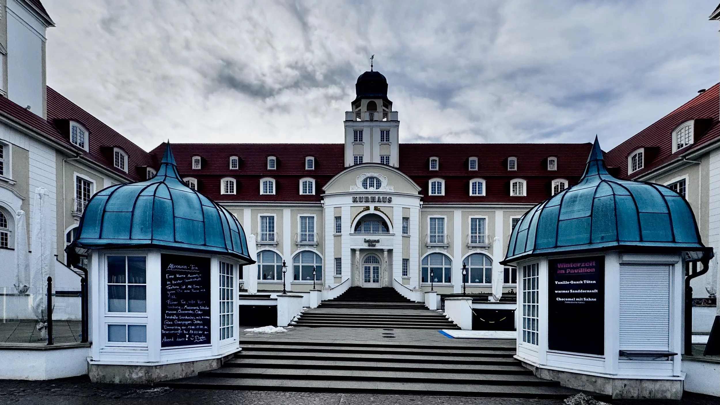 Ein prächtiges, historisches Gebäude mit einem Schriftzug "Kurhaus" über dem Eingang, umgeben von zwei Pavillons mit blauen Dächern, bei bewölktem Himmel.