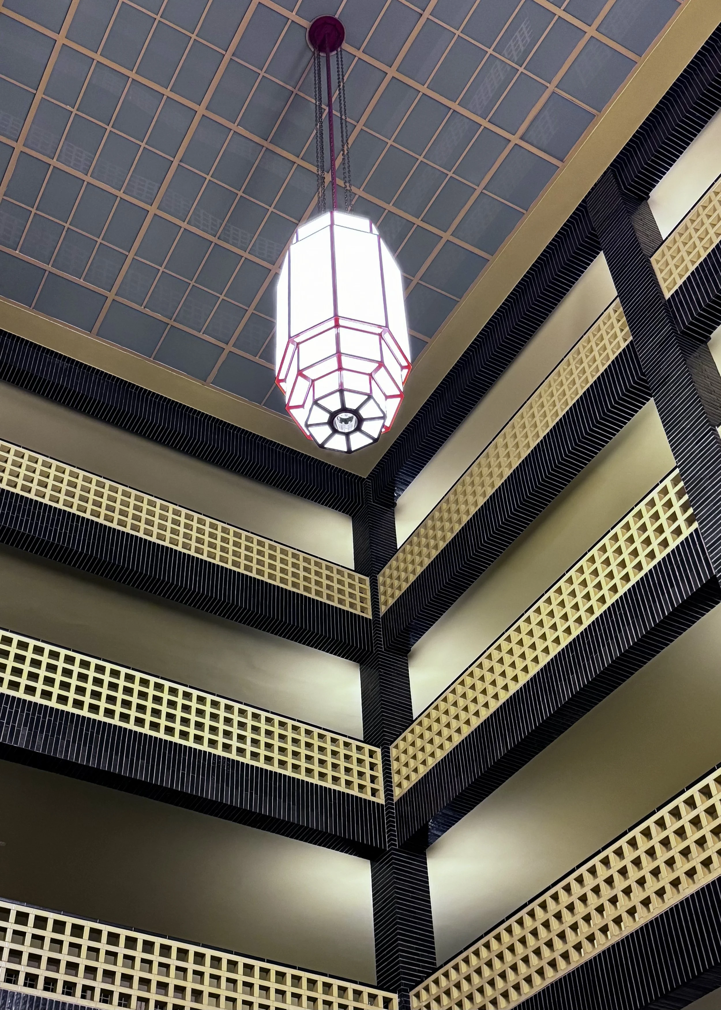 Innenaufnahme eines Gebäudes mit mehreren Etagen, einem geometrischen Gemälde an der Decke und einer auffälligen geometrischen Hängeleuchte.