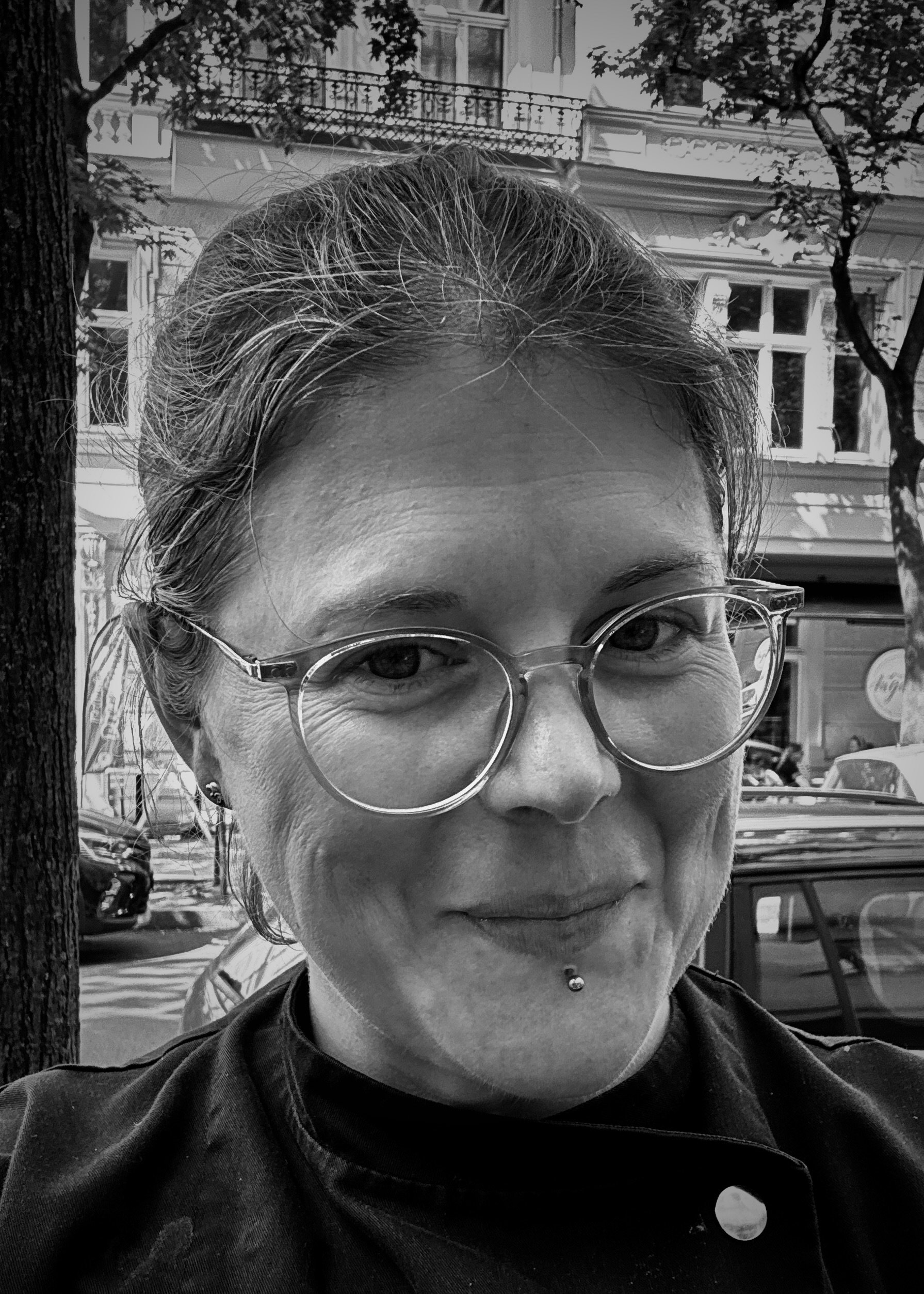 Schwarz-weiß-Foto einer Frau mit Brille und Piercing im unteren Lippenbereich, auf einer Straße mit Bäumen und Gebäuden im Hintergrund.