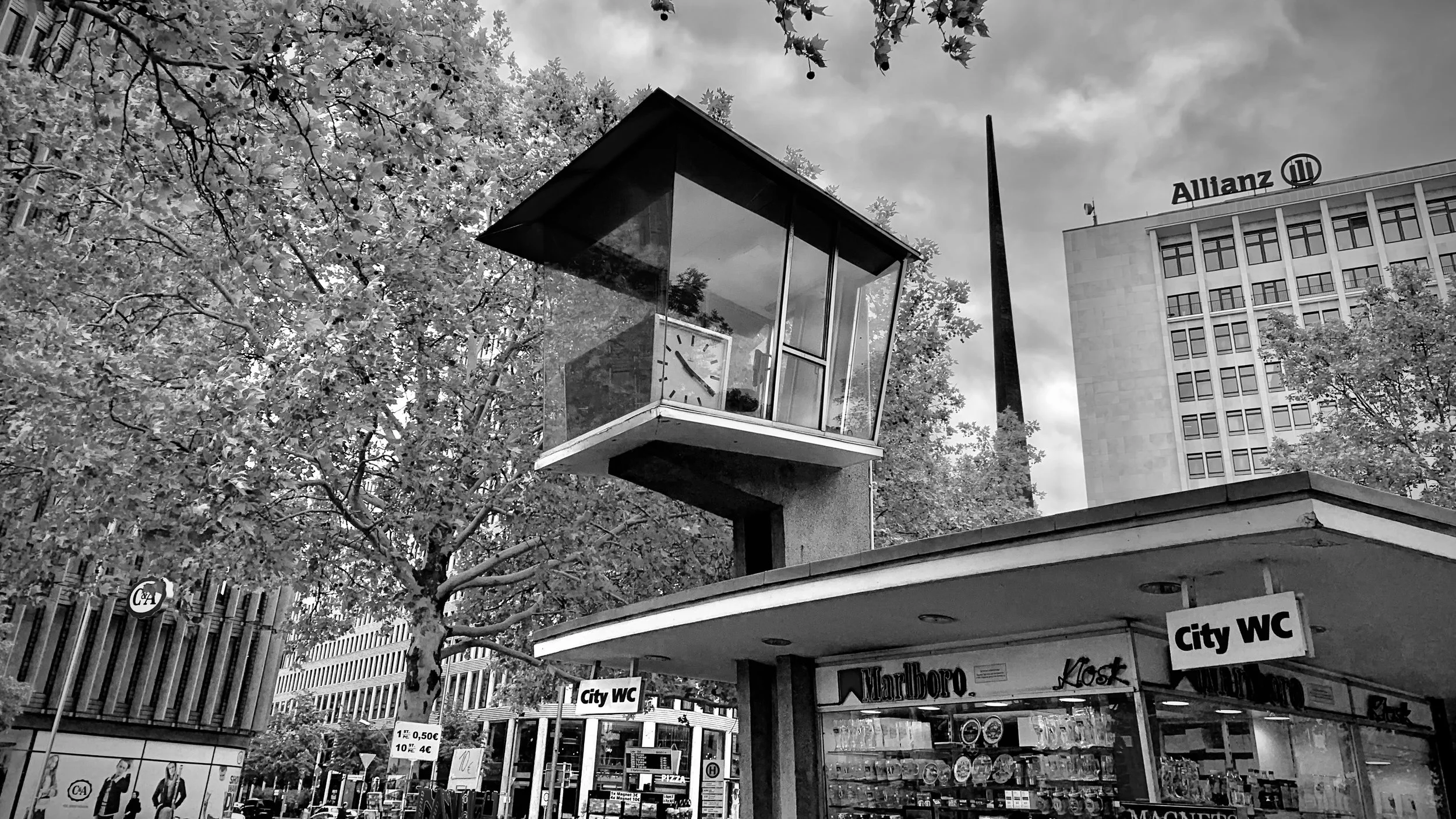 Schwarz-weiße Aufnahme eines öffentlichen Platzes mit einem modernen Gebäude im Hintergrund, einem überdachten Kiosk im Vordergrund, umgeben von Bäumen und Werbeschildern.