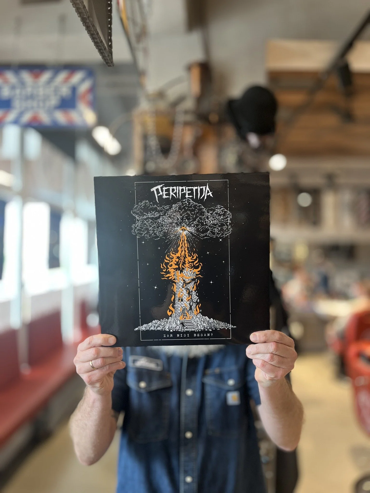 A person holding a black booklet with a graphic design of a mushroom cloud and a guitar, with the words 'PERIPETIJA' and 'ZAR NİSİ BESAN?' printed on it, in a bookstore or library setting.
