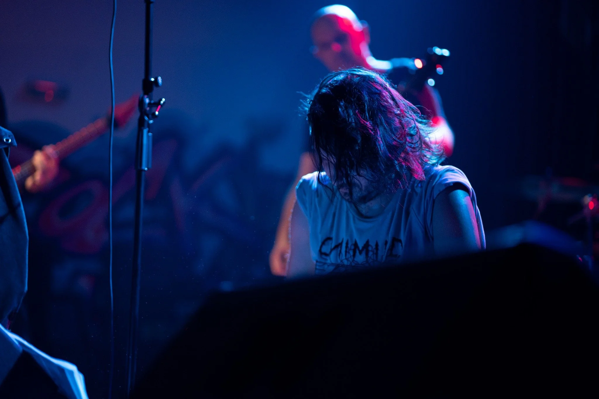A woman with wet hair performing on stage, with a band member playing guitar in the background, illuminated by blue and pink stage lights.