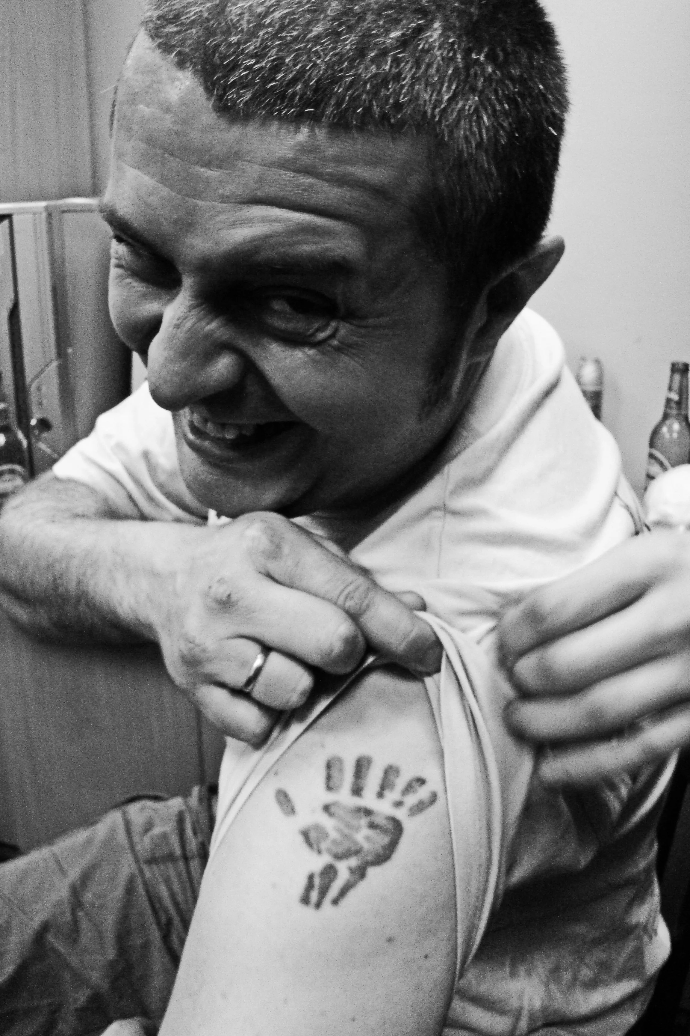 A young man with a buzz cut hairstyle, smiling and looking down, wearing a white shirt, and showing a small tattoo of a handprint on his forearm.