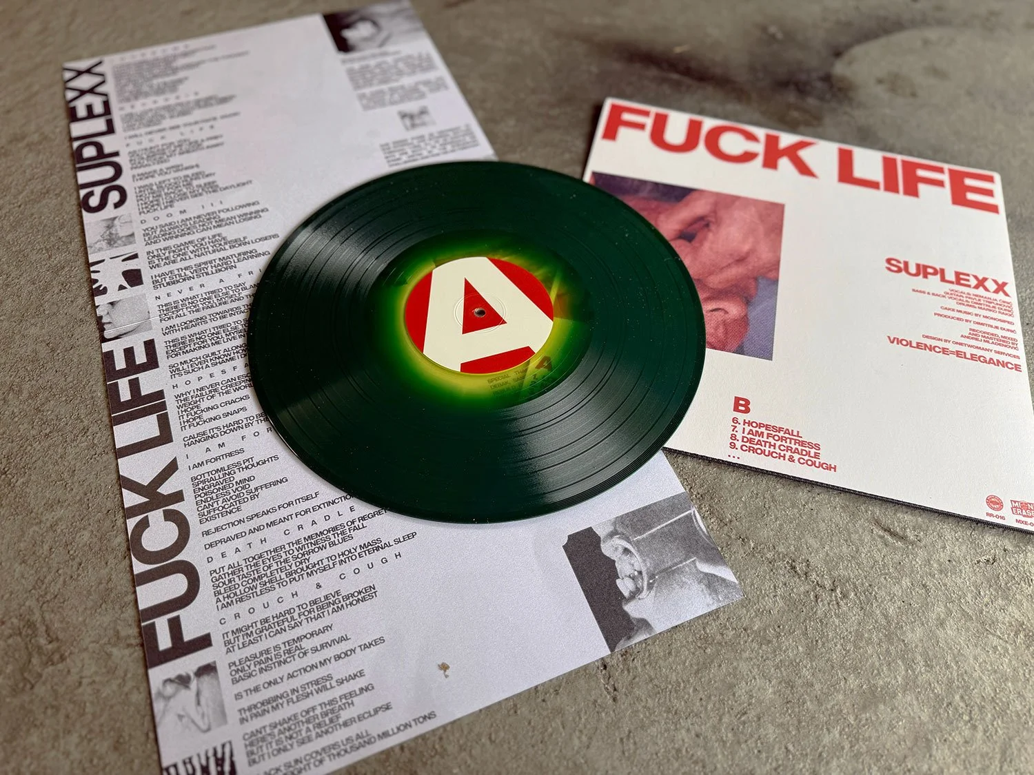A vinyl record with a red and white label resting on printed papers, with another printed paper beside it featuring bold red text.