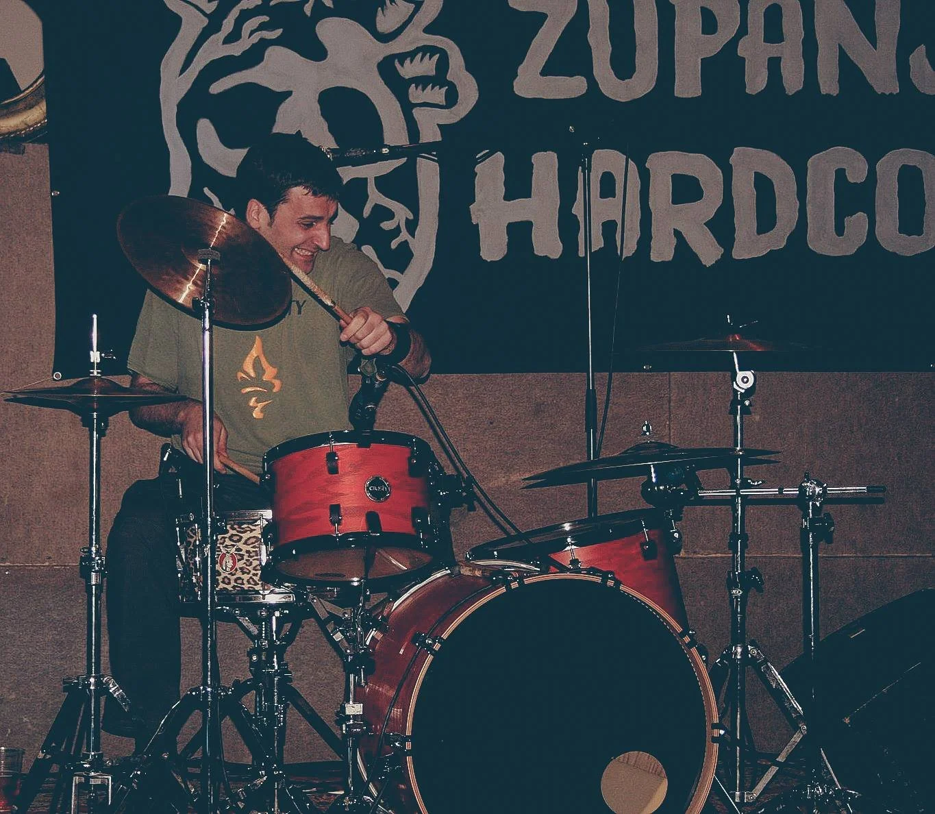 A young man playing a red drum set on stage, smiling, with a backdrop that reads 'ZUPAN'S HARDCOCK' and features a skeleton graphic.