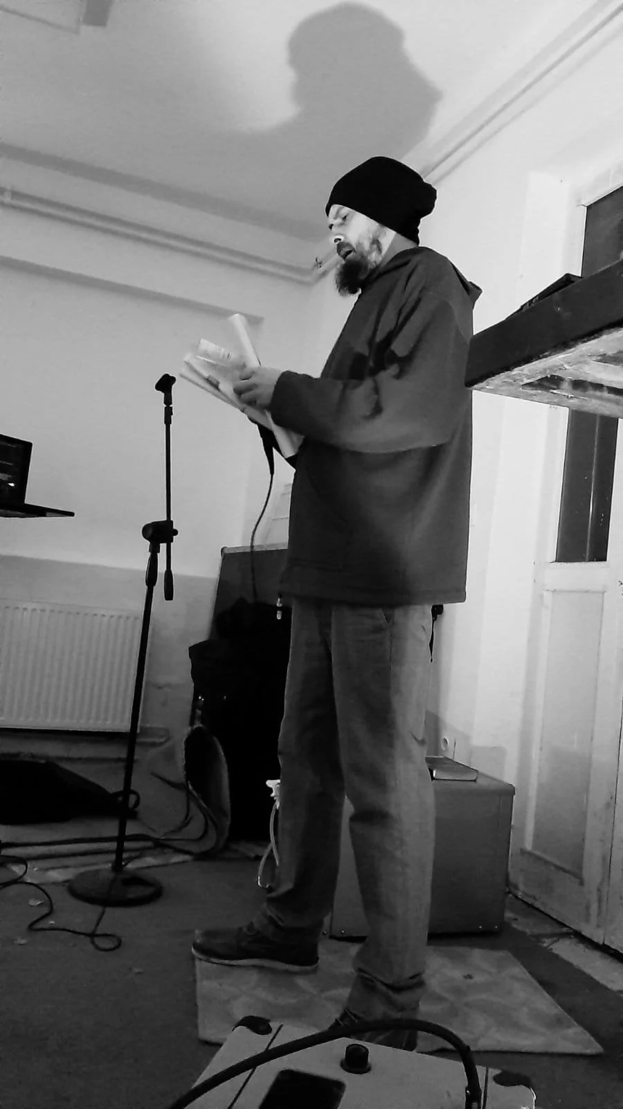 A man with a beard, wearing a beanie and casual clothes, standing on a small platform reading from a paper in a room with equipment, microphone, and a laptop.