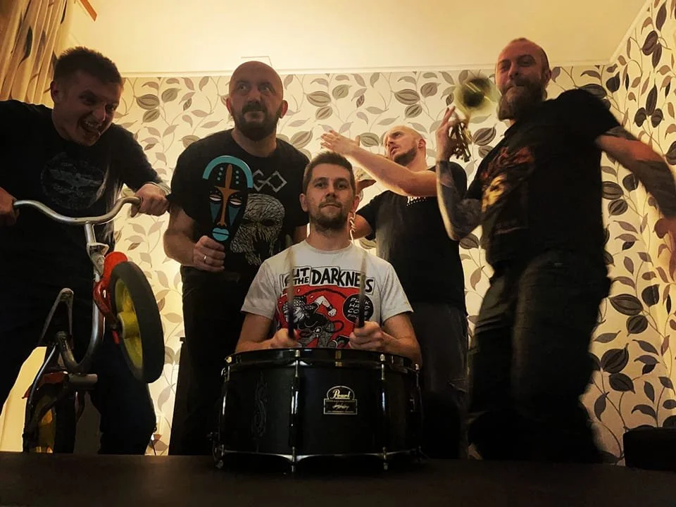 Five men in a room with patterned wallpaper, some holding musical instruments and others raising their arms or posing.