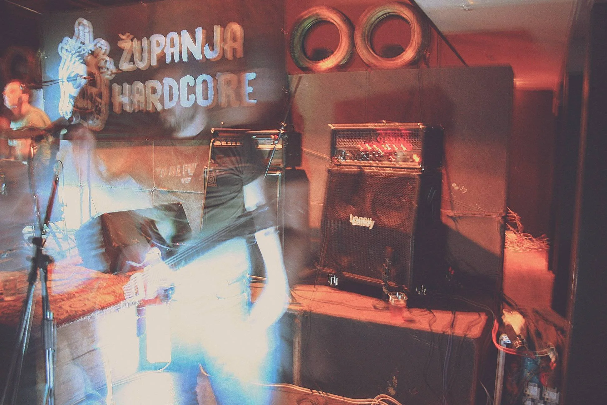 A stage with musical equipment including a guitar amplifier, speakers, and microphones, with a colorful sign that reads 'ZUPANJA HARDCORE' on the wall.