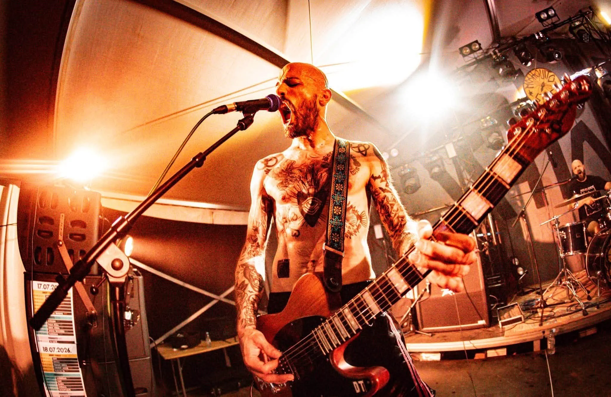 A shirtless male musician covered in tattoos passionately singing into a microphone while playing an acoustic guitar during a live performance. The background shows stage lighting and sound equipment.