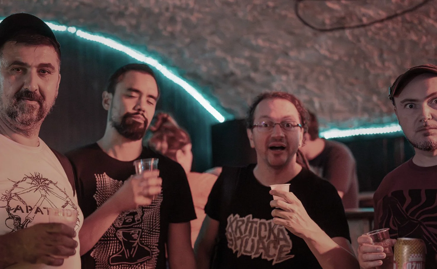 Four men at a bar or club with neon lighting, holding drinks, with a rough stone ceiling overhead.